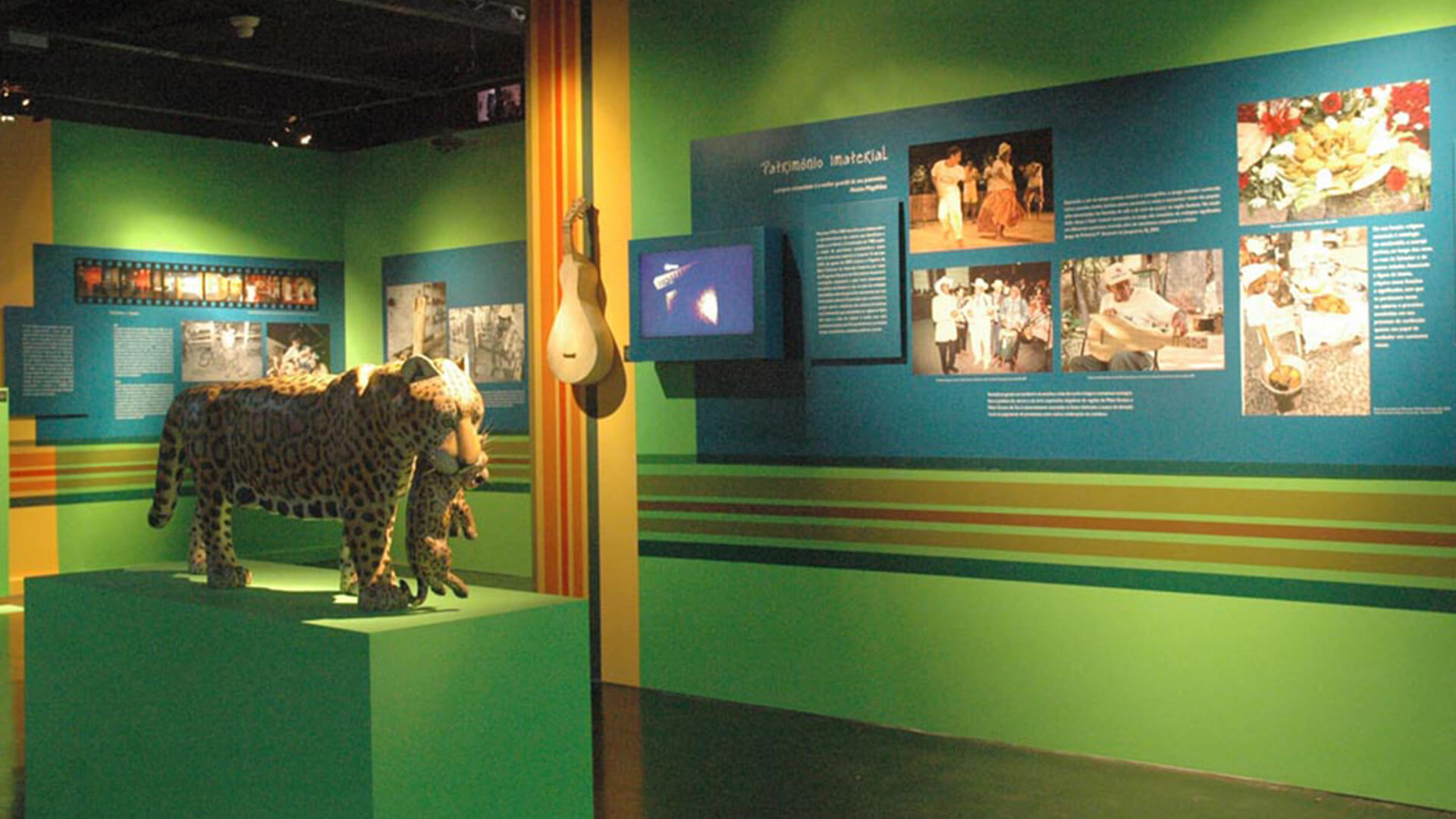 Ambiente de exposição com paredes em tons de verde, azul e faixas horizontais coloridas. À esquerda, uma escultura de uma onça-pintada carregando um filhote pela boca, sob um pedestal verde. Na parede lateral, um painel azul apresenta textos, uma pequena tela e uma sequência de fotos coloridas de danças e comidas.