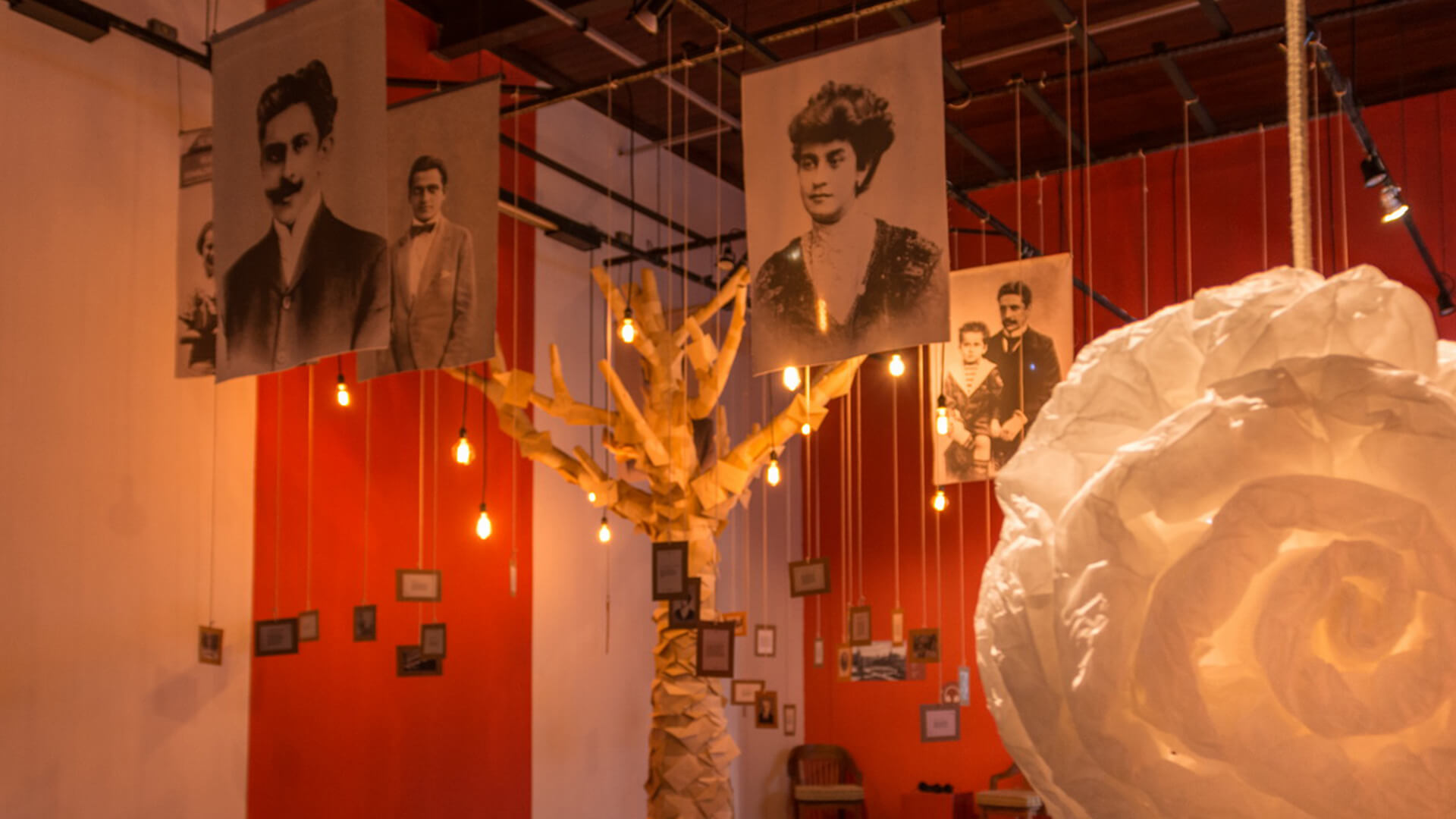 Instalação de arte com fundo vermelho, exibindo retratos históricos em preto e branco pendurados do teto. Uma escultura de tronco de árvore, com pequenas molduras penduradas, fica no centro, iluminada por lâmpadas suspensas. Uma grande flor branca de papel está à direita.