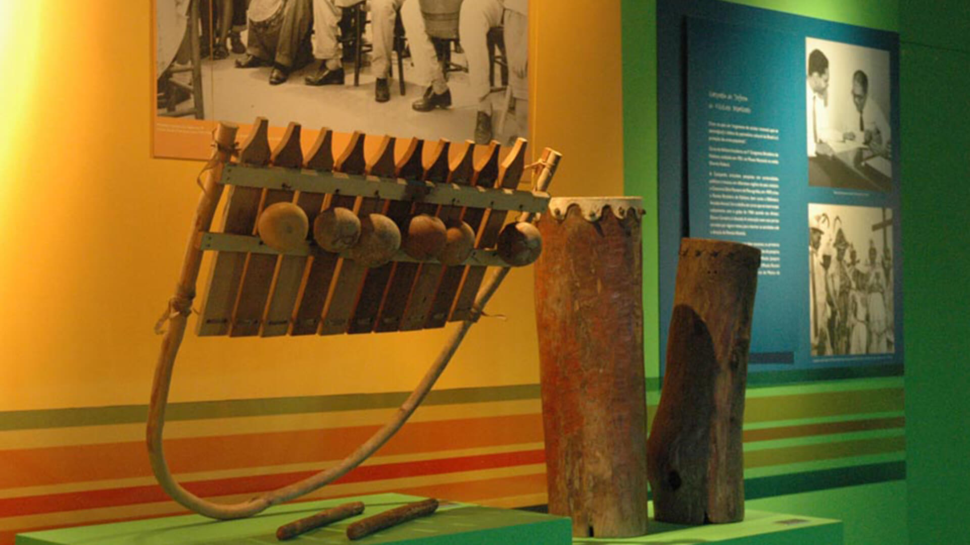 Paredes amarelas e verdes. Em primeiro plano, há um instrumento musical de percussão feito de madeira. Ao lado, há dois troncos de madeira que formam tambores, com pedaço de lona ou couro cobrindo o topo e preso nas lateriais. No fundo, um painel azul com texto e fotos históricas.