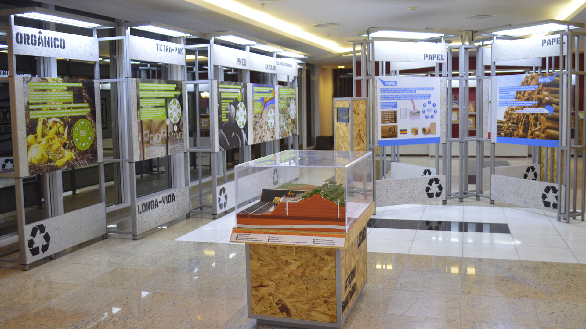 Visão ampla de um estande de exposição sobre reciclagem. O espaço é preenchido com vários painéis informativos suspensos (rotulados ORGÂNICO, TETRA-PAK, PNEU, PAPEL etc.) e no centro há um modelo de exibição em escala (Lixão vs. Aterro Sanitário) dentro de uma caixa de acrílico. Símbolos de reciclagem estão visíveis na base das estruturas.