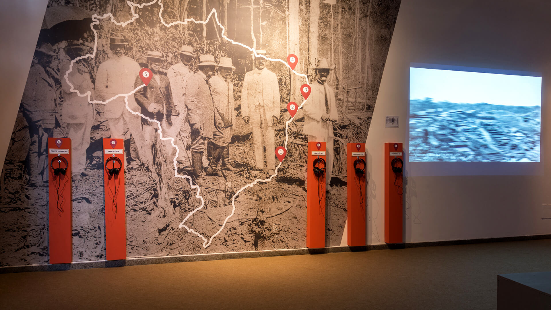 Exposição de museu com uma grande parede coberta por uma fotografia histórica em preto e branco de um grupo de homens em um ambiente rural, sobreposta por um mapa de contorno branco com pinos vermelhos. Seis painéis estreitos e laranja, cada um com fones de ouvido pendurados, estão alinhados em frente ao mural.