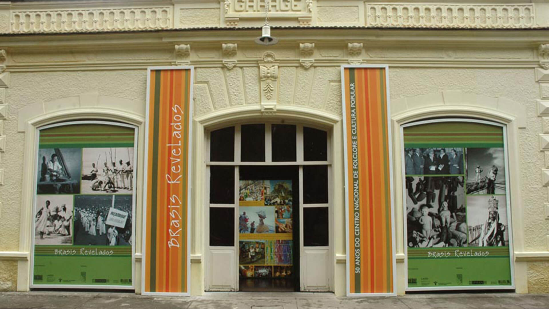Fachada de um prédio histórico de cor creme. As janela laterais são cobertas por grandes painéis verdes com uma colagem de fotos históricas em preto e branco. Duas faixas verticais coloridas (laranja, verde e amarelo) entre as janelas e a porta central, com o texto BRASIS REVELADOS e 50 ANOS DO CENTRO NACIONAL DE FOLCLORE E CULTURA POPULAR.