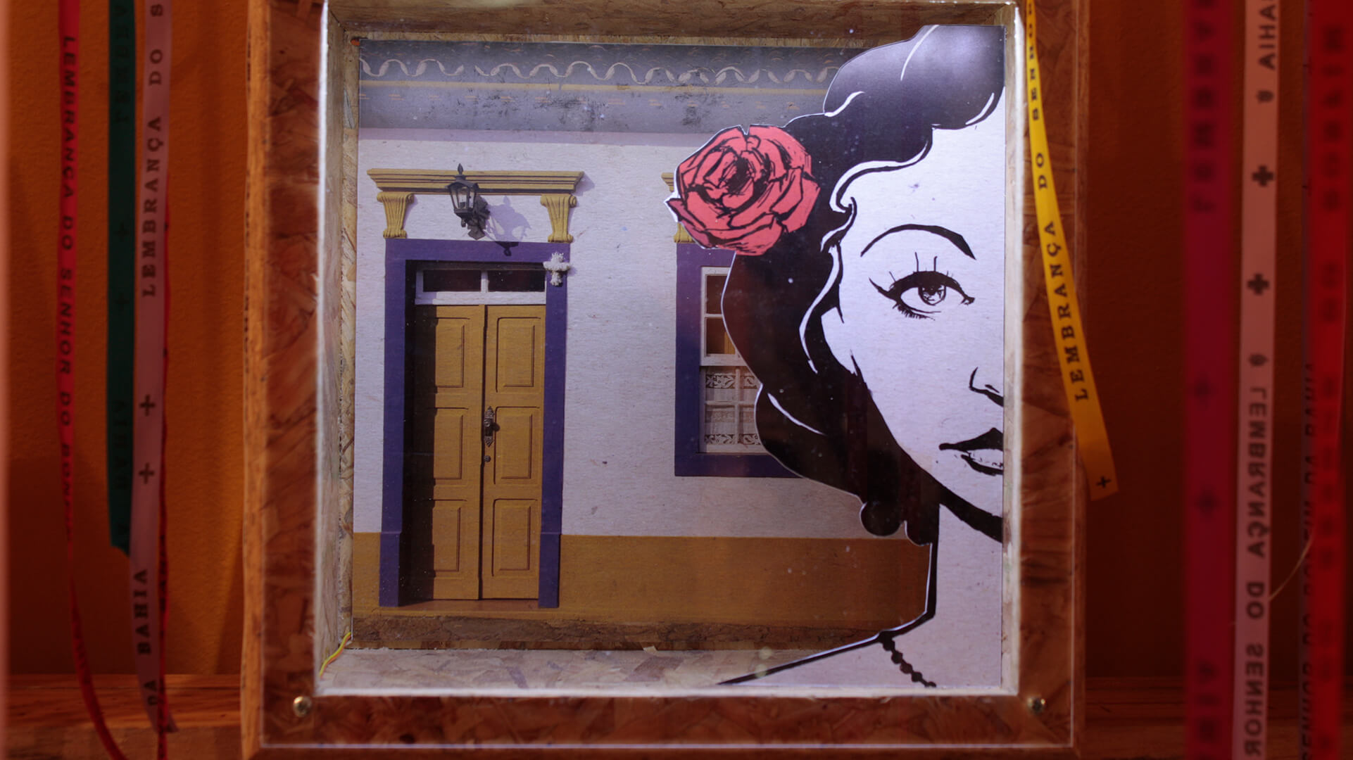 Cena de arte em caixa (diorama) com uma ilustração de rosto feminino de lado e uma rosa vermelha no cabelo, sobreposta a um fundo que simula a fachada de uma casa com porta amarela e batente roxo, emoldurada por madeira e fitas de souvenir.