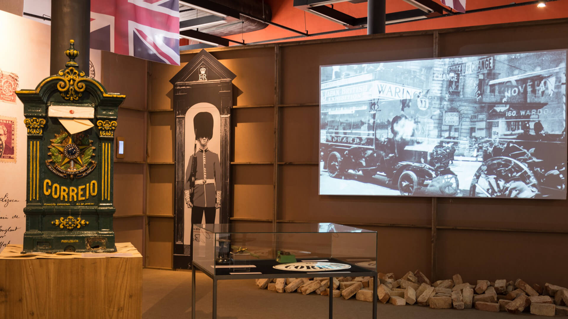 Exposição de museu com um marco de correio antigo verde e dourado à esquerda, uma silhueta em escala real de um guarda real britânico no centro, e uma projeção em preto e branco de uma cena de rua histórica em uma grande tela, com bandeiras do Reino Unido no topo.