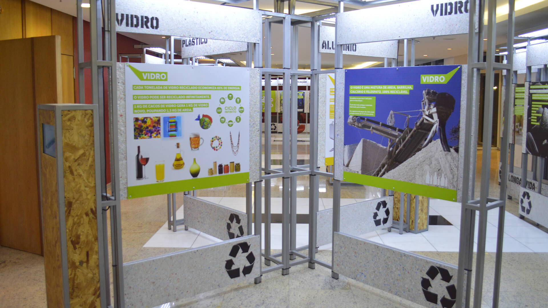 Stand de exposição sobre reciclagem, com painéis informativos suspensos por uma estrutura de metal. Painéis centrais grandes detalham a reciclagem de VIDRO, incluindo gráficos de produtos e uma foto de uma máquina de reciclagem. Símbolos de reciclagem pretos estão na base do estande.