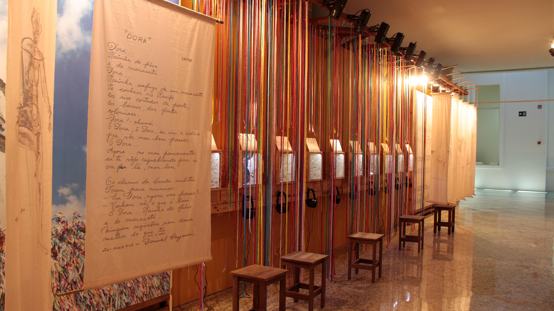 Instalação artística em corredor, com uma parede com longas fitas coloridas penduradas, nichos iluminados e cada um com um fone de ouvido pendurado, e um grande banner de tecido em primeiro plano com um texto escrito à mão (poema 