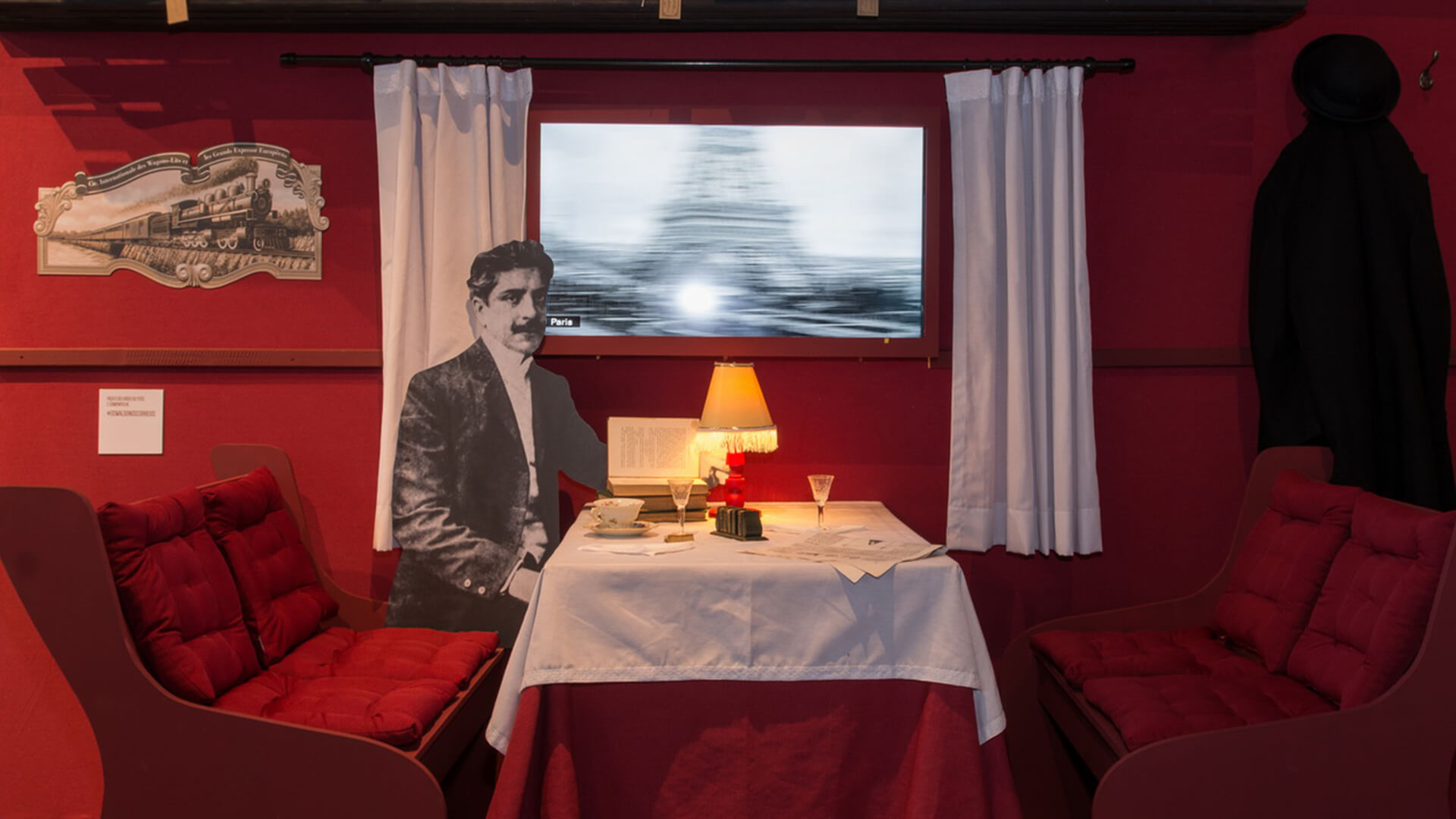 Cenografia simulando o interior de um vagão de trem em vermelho intenso, com dois assentos estofados vermelhos, uma mesa posta, um recorte em tamanho real de Oswaldo Cruz sentado e uma tela simulando o movimento através de uma janela que mostra a Torre Eiffel.