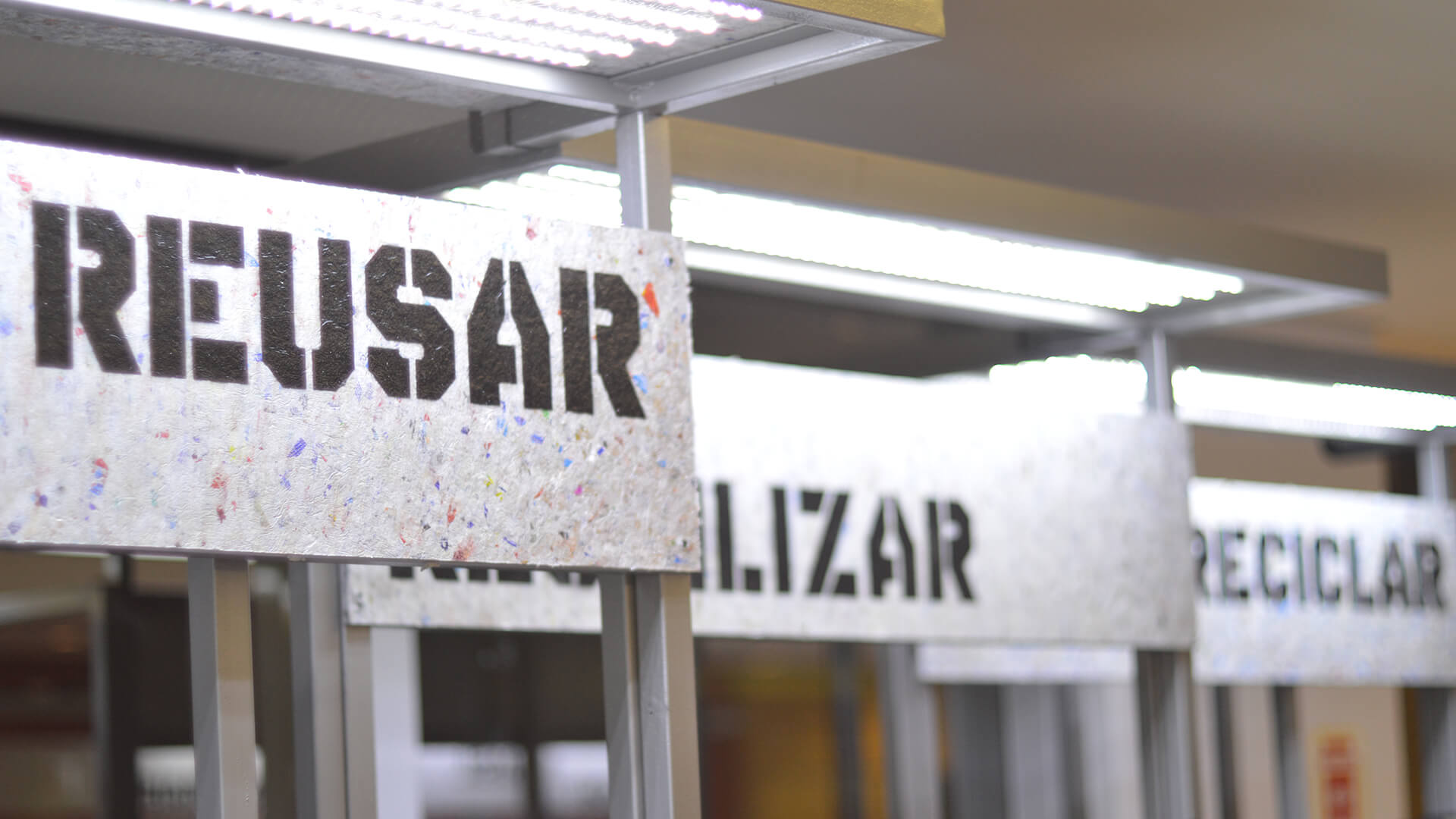 Placas de sinalização verticais, feitas de material reciclado com pontos coloridos, exibindo as palavras da política dos 3Rs: REUSAR, REUTILIZAR e RECICLAR (bem ao fundo, parcialmente visível).