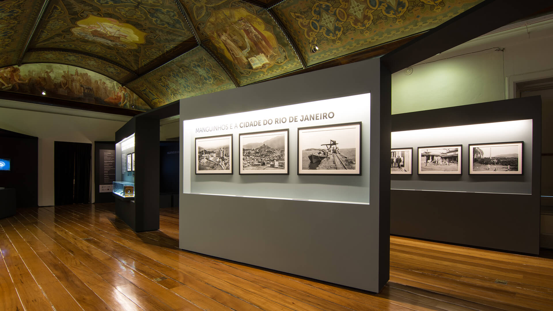 Interior de galeria com piso de madeira polida, teto decorado com afrescos, e uma exposi&ccedil;&atilde;o com um grande painel intitulado 