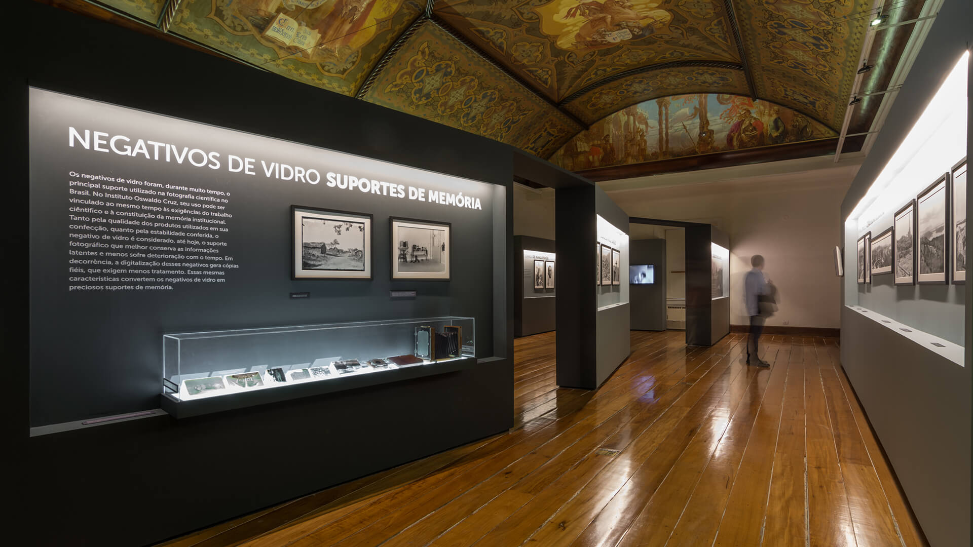 Interior de galeria com piso de madeira polida, teto decorado com afrescos, e uma exposição com um grande painel intitulado 