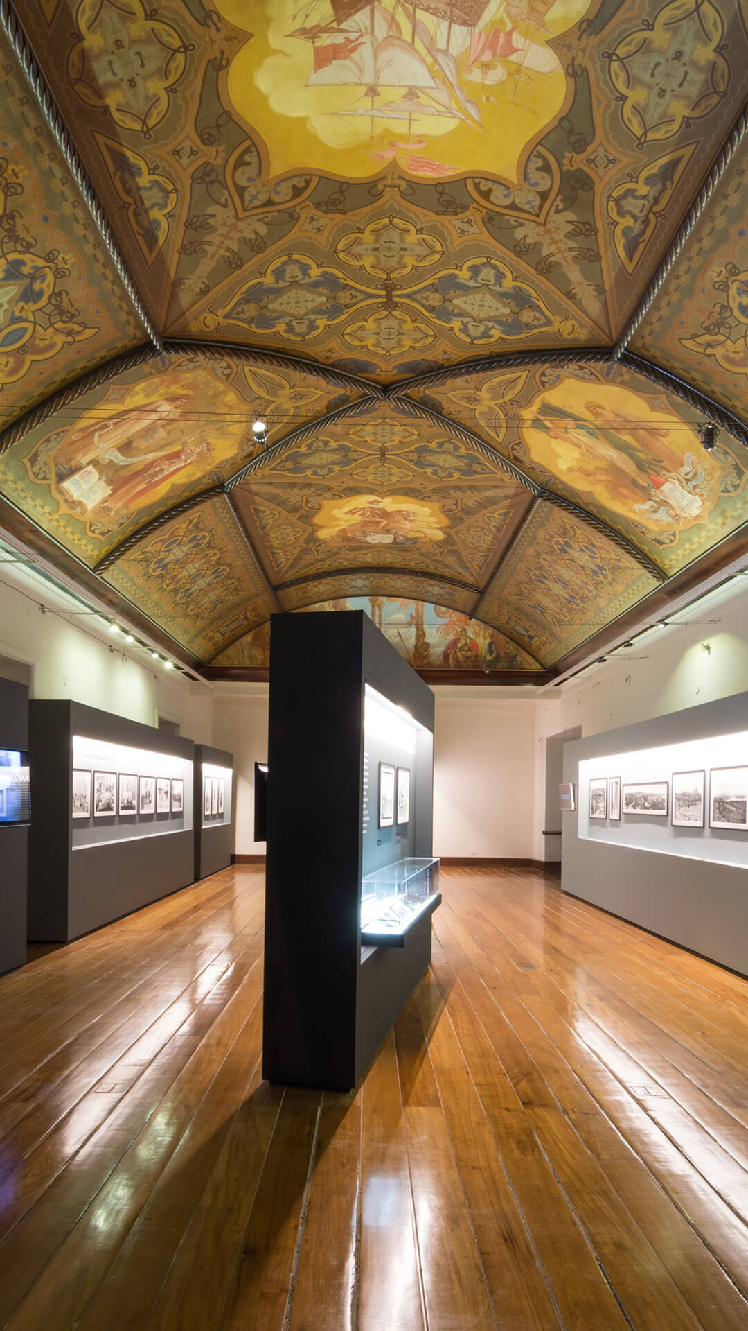  Interior de uma galeria com piso de madeira polida, um teto pintado com afrescos e arcos decorados, e exposições de fotografias em preto e branco em painéis cinzas e pretos