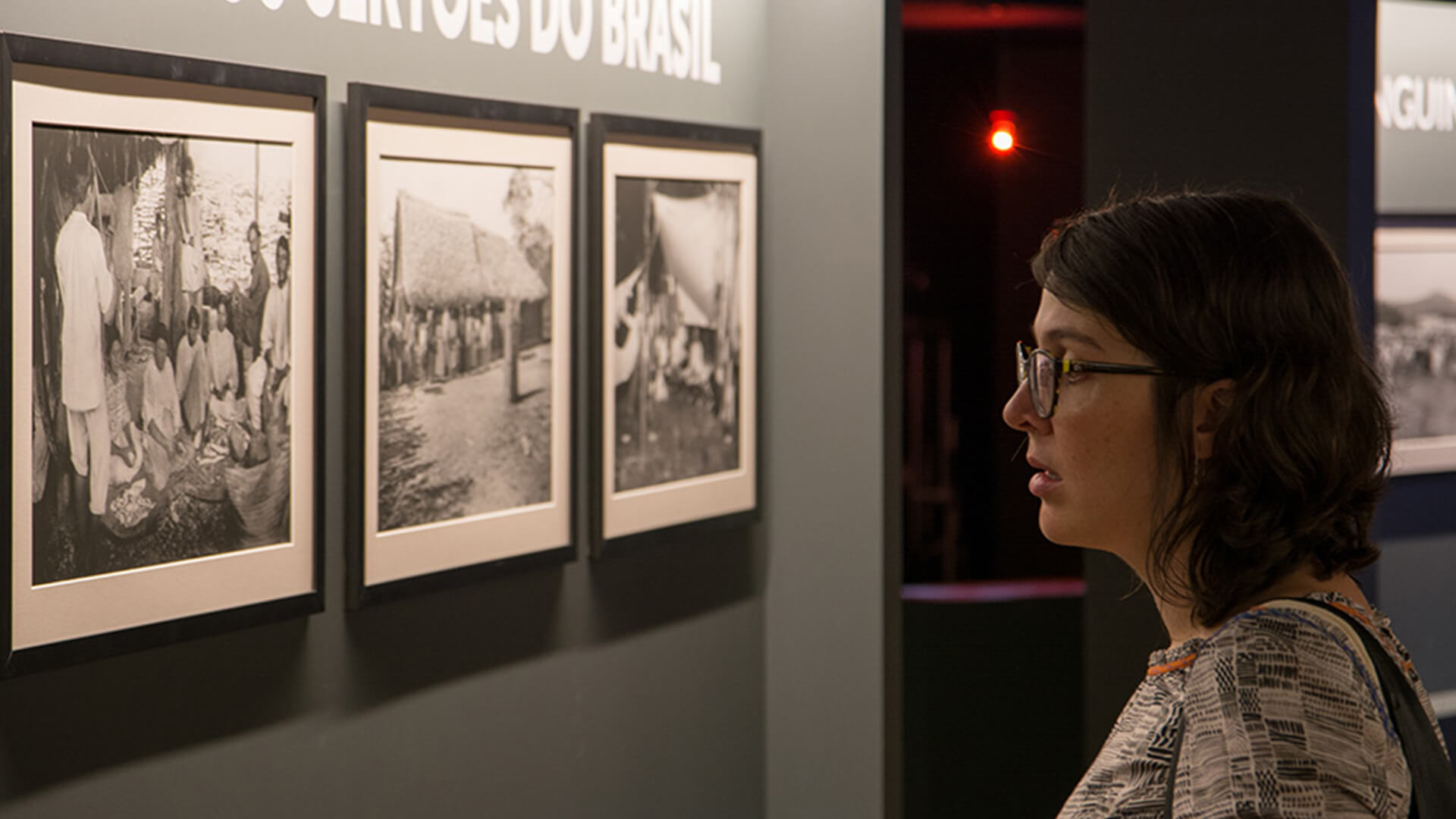 Close de mulher observando três quadros de fotografias em preto e branco, com moldura preta, sob parede cinza. A mulher é branca, tem cabelos curtos, lisos e escuros. Ela usa óculos e está de perfil para a foto.