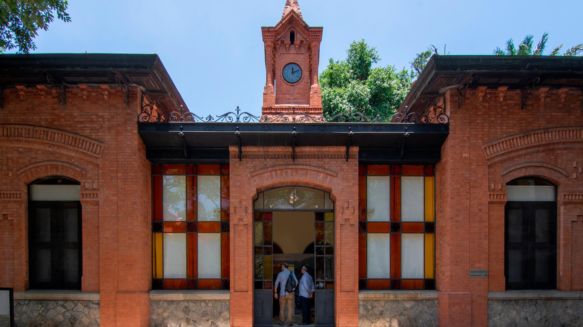 Fachada de um edifício histórico de tijolos vermelhos com telhado de ferro e uma torre central com relógio. A entrada principal tem um arco e grandes janelas retangulares com vitrais em cores como amarelo, vermelho e branco, e duas pessoas estão entrando.