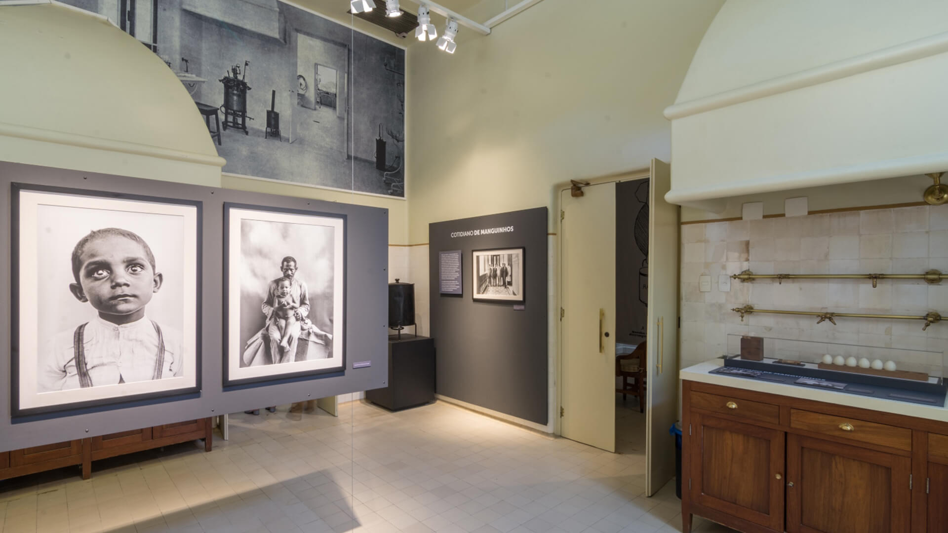 Interior com paredes de cor creme, piso de ladrilhos e mobiliário de madeira escura com uma coifa embutida. A exposição apresenta fotografias em preto e branco, incluindo retratos grandes de crianças e uma foto maior no alto da parede, mostrando um interior antigo.