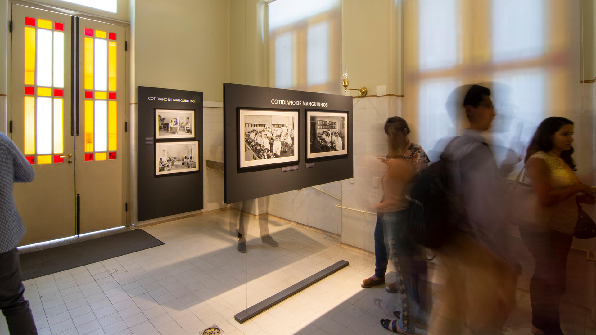Chão de ladrilhos e uma porta com vidros coloridos (amarelo e vermelho). Um painel de exposição escuro com o título 