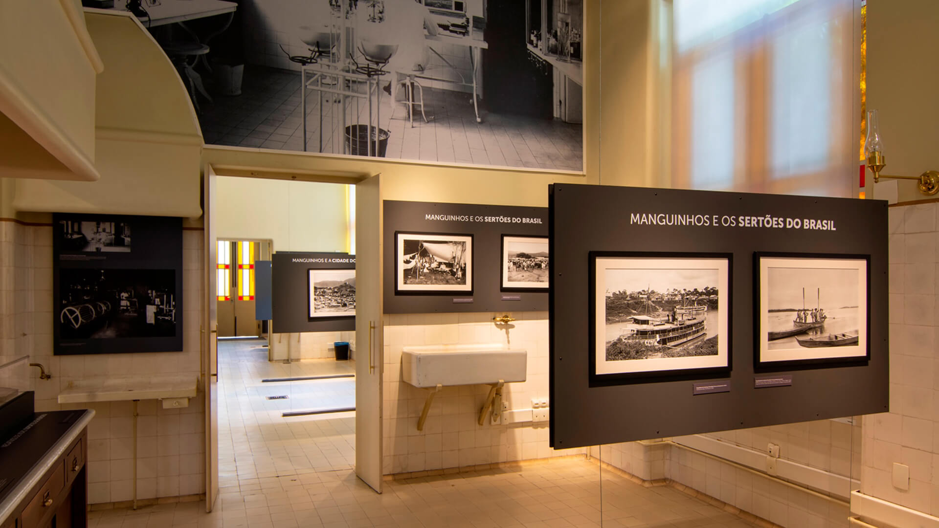 Interior de um espaço de exposição que parece ser um laboratório ou cozinha antiga adaptada, com azulejos brancos nas paredes, uma pia de porcelana e pisos de ladrilhos claros. A exposição apresenta painéis escuros com o título 