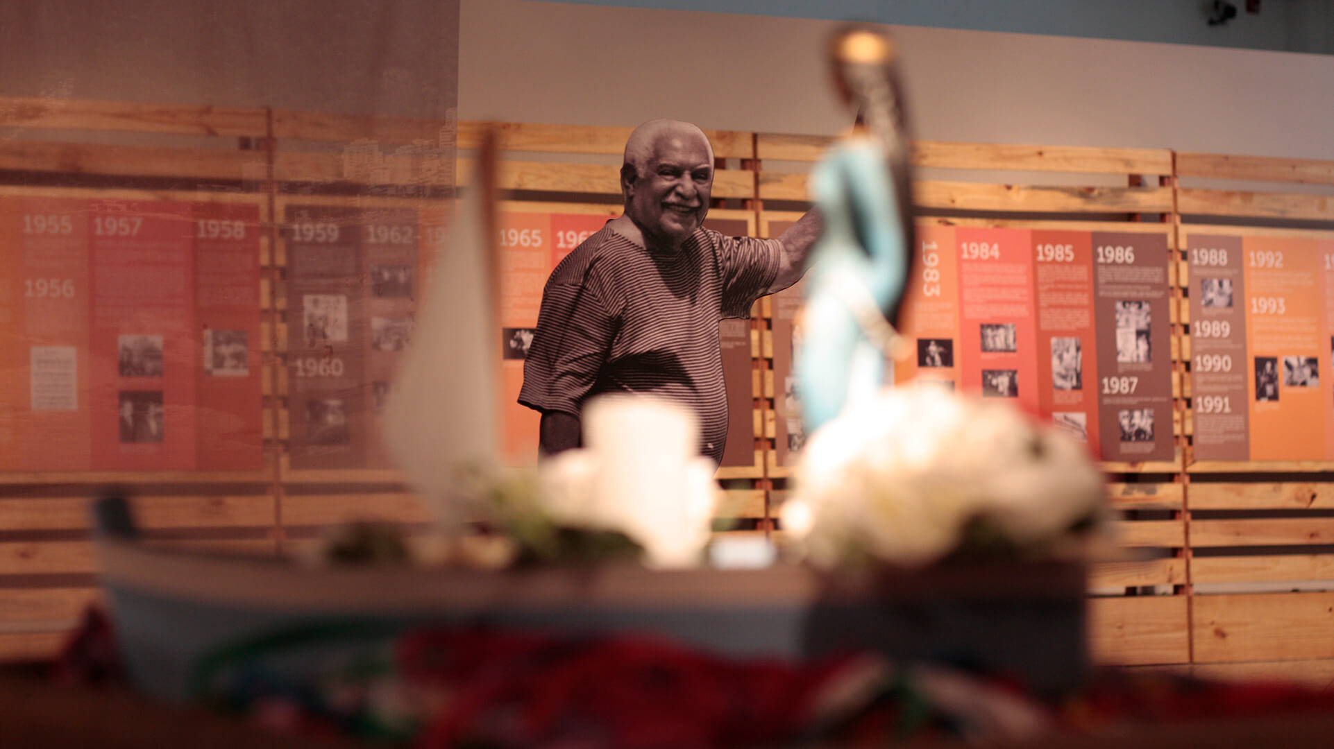 Imagem de exposi&ccedil;&atilde;o focada  com uma foto grande em s&eacute;pia de um homem sorrindo (possivelmente Dorival Caymmi), e em primeiro plano, desfocado, um barco de oferendas com uma est&aacute;tua e flores.
