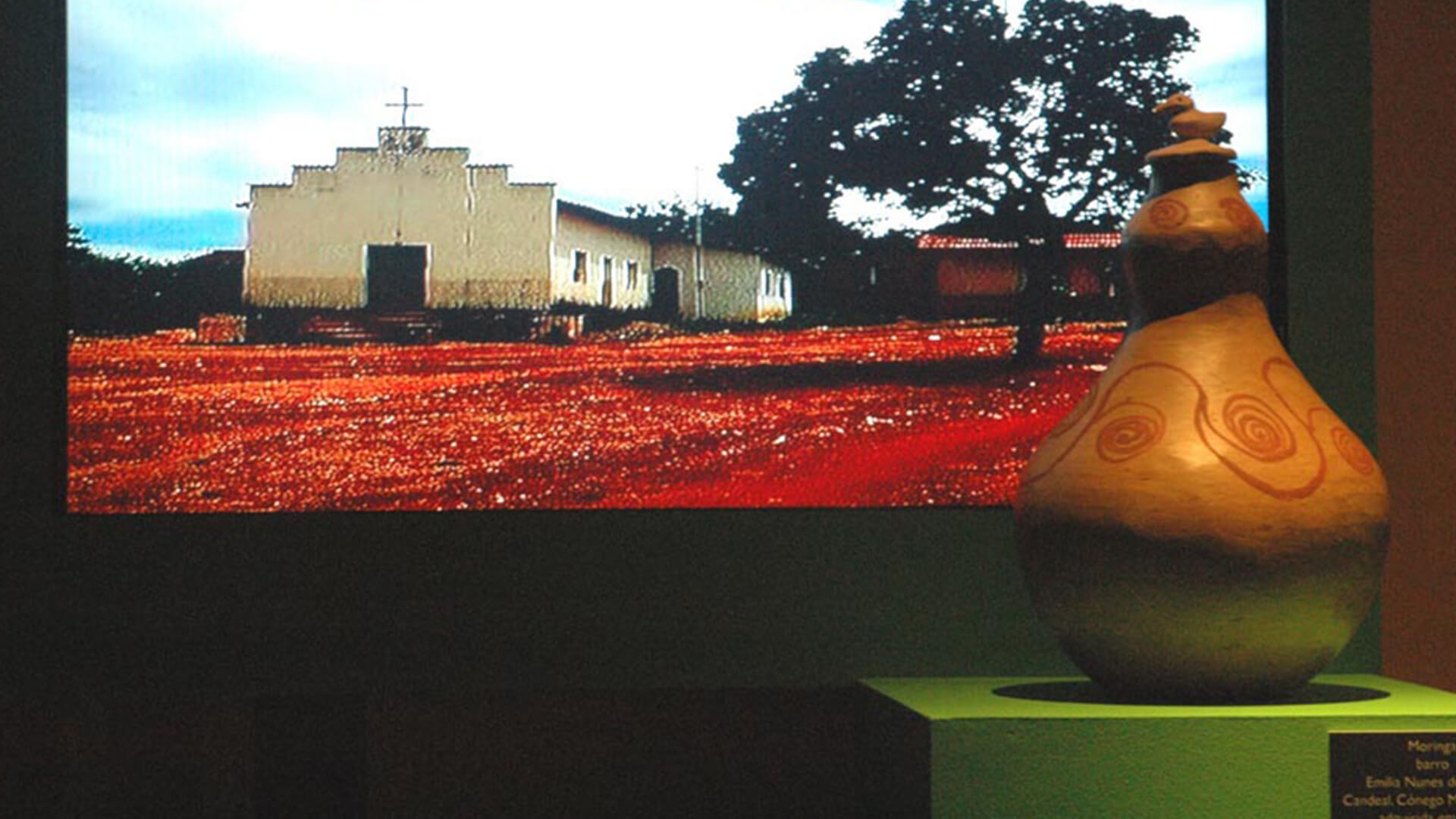 Uma grande tela exibe uma fotografia de uma igreja branca simples com uma cruz no telhado, em um solo de terra vermelha, com uma grande &aacute;rvore &agrave; direita. Em primeiro plano, no canto inferior direito, h&aacute; uma pe&ccedil;a de cer&acirc;mica marrom em forma de caba&ccedil;a, com desenhos em espiral em cor vermelha, sobre um pedestal verde.