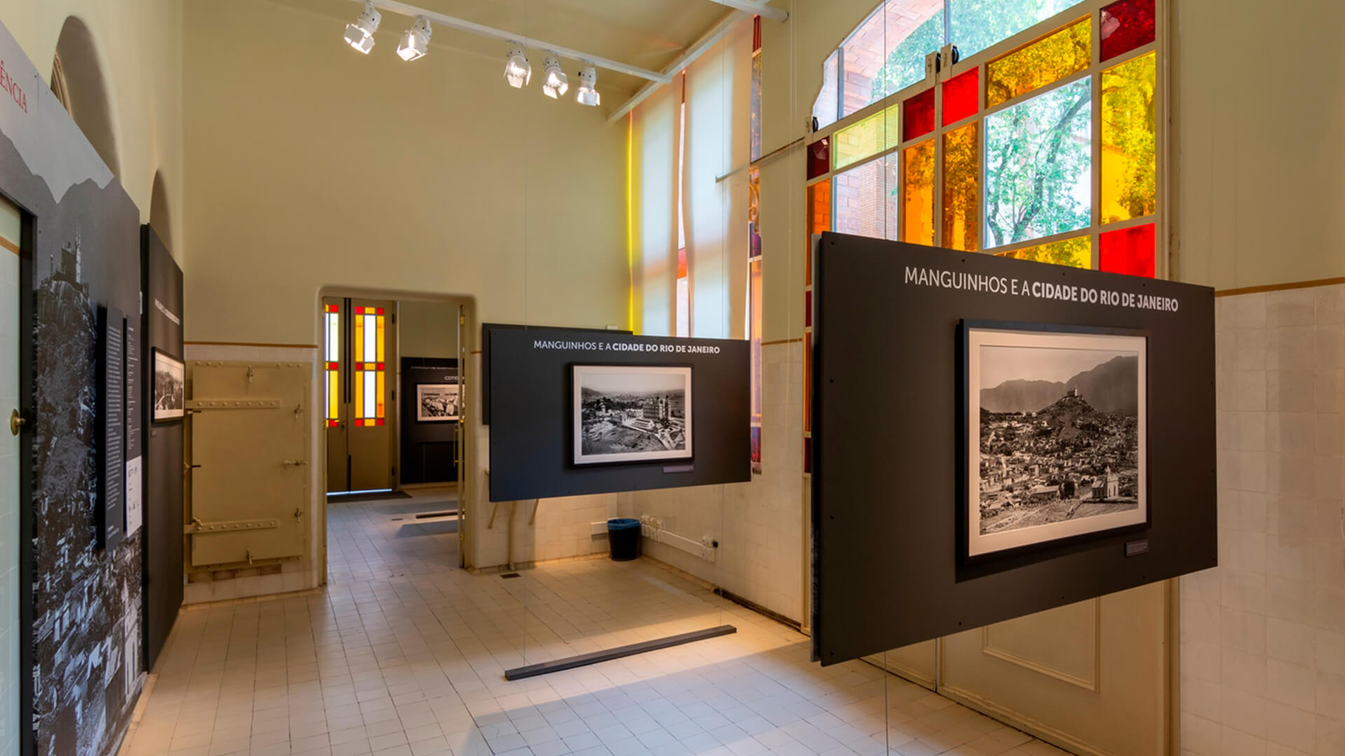 Interior de uma sala de exposi&ccedil;&atilde;o de museu com paredes de cor clara, piso de ladrilhos e janelas grandes com vidros coloridos (vermelho, amarelo e branco) que iluminam o ambiente. Pain&eacute;is escuros flutuantes exibem o t&iacute;tulo 