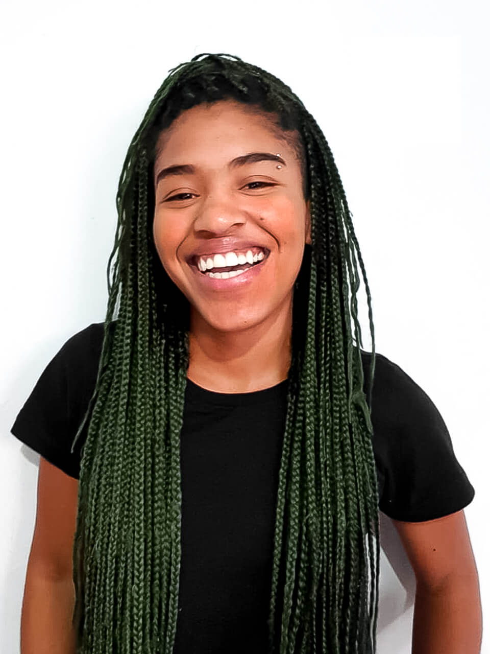 Mayara é negra, usa tranças box brainds verde e uma camisa preta de manga curta.