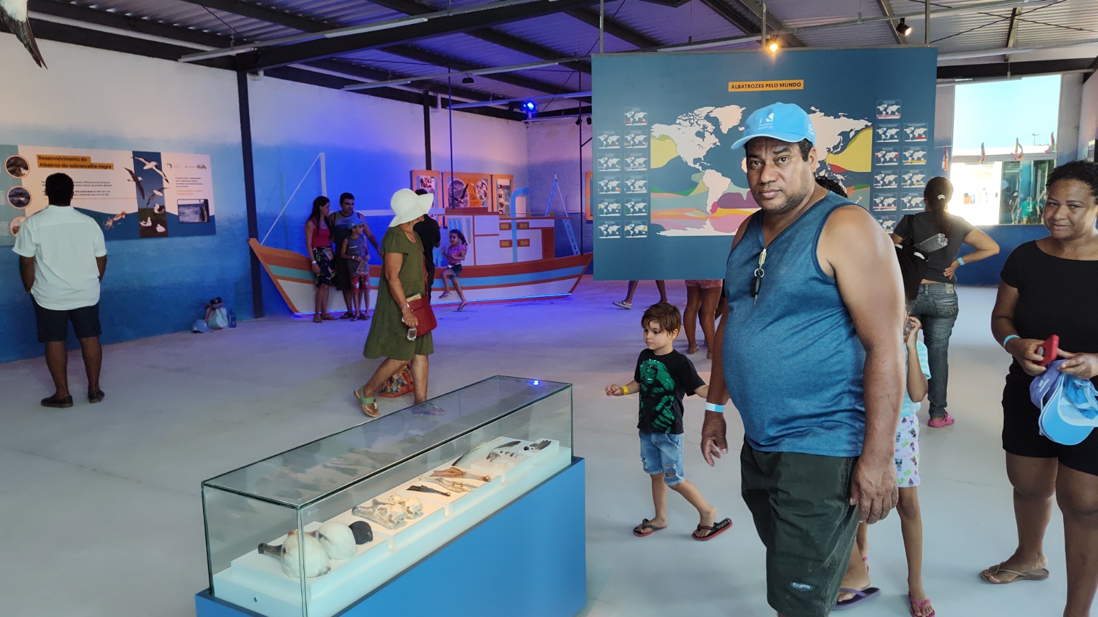 Visitantes circulam pela exposição Albatroz, que inclui uma vitrine com objetos, mapa-mundi informativo pendurado no centro da sala e ao fundo uma réplica de um barco de pesca.