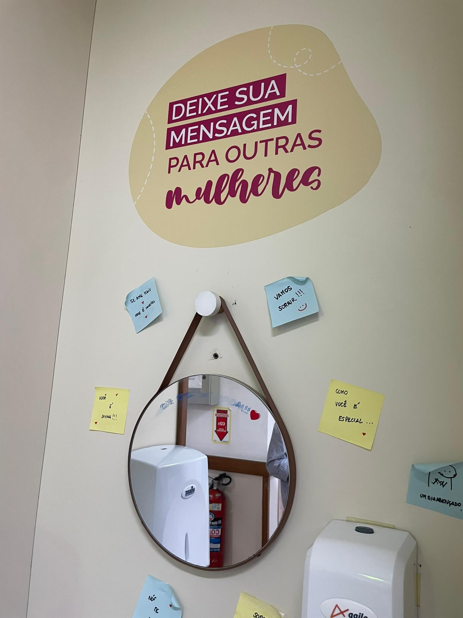 Área para lavar as mãos, com paredes em tom de amarelo claro, espelho redondo pendurado em suporte, acima, um adesivo com a frase 