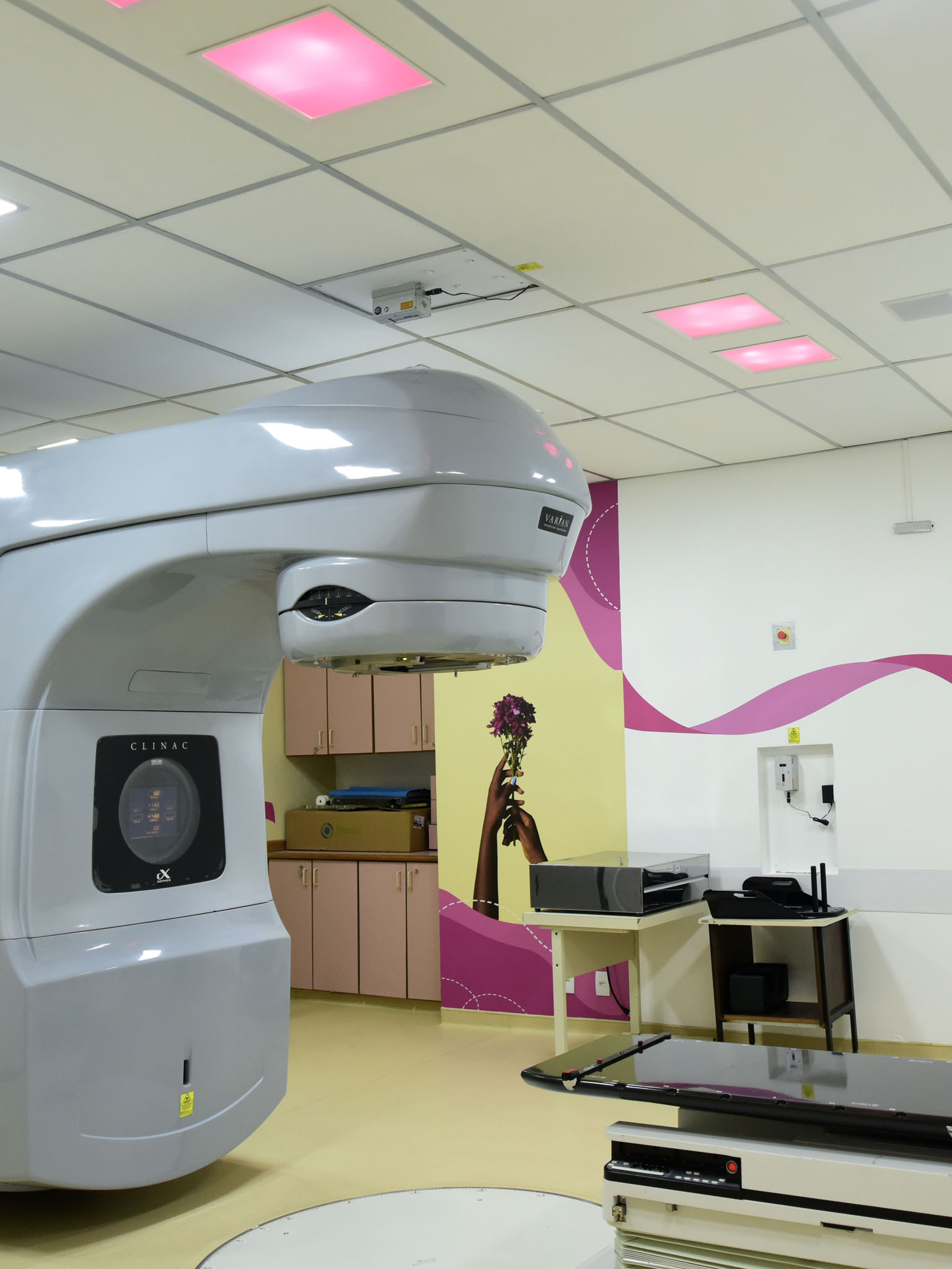 Sala de tratamento de câncer com um grande equipamento de tratamentos dos pacientes no centro, tetos com luzes rosas, e um mural artístico na parede, sugerindo um ambiente de tratamento humanizado.