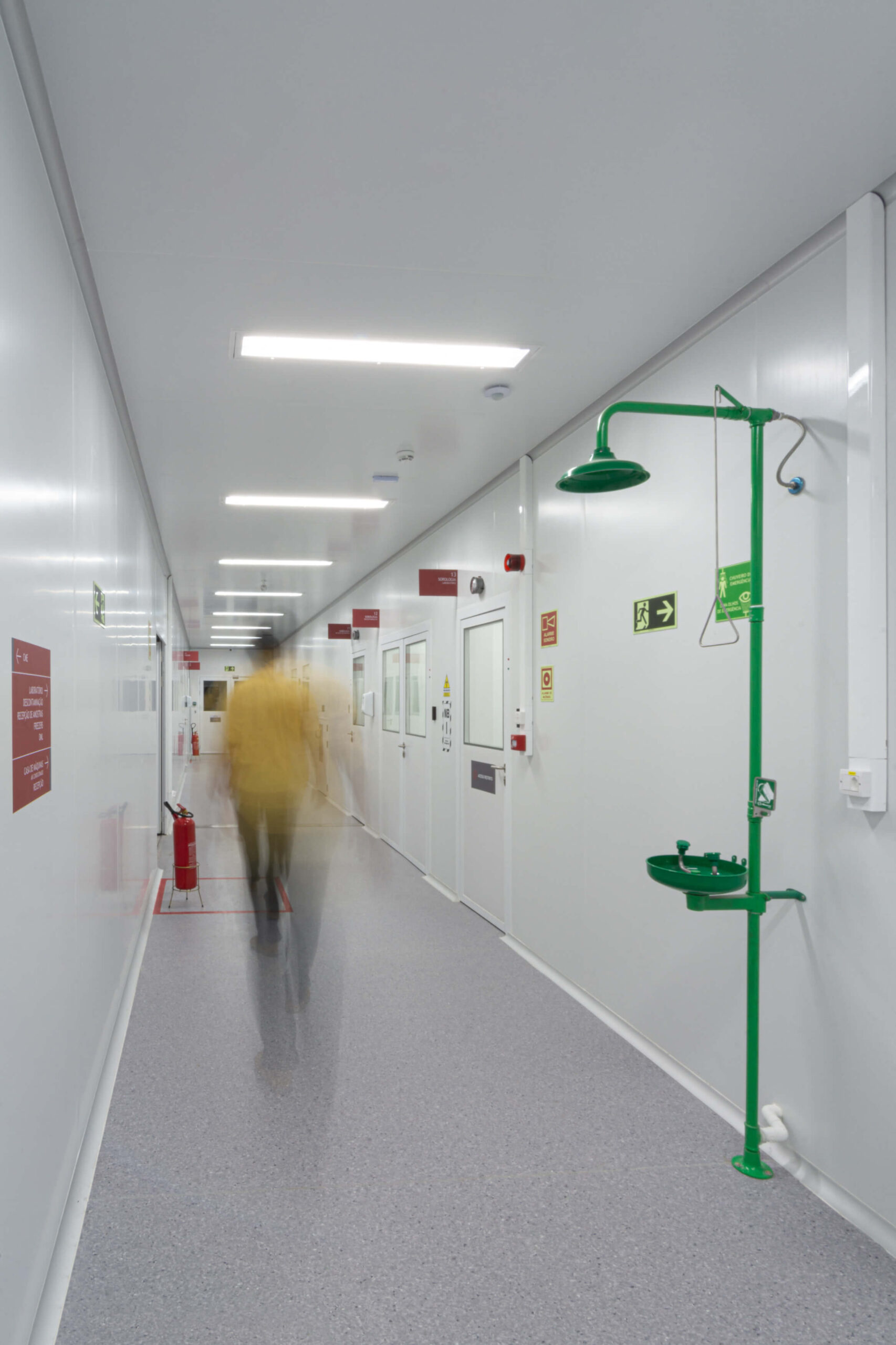 Corredor de laboratório com paredes e portas brancas, placas vermelhas de identificação numeradas.  Um pessoa caminha pelo corredor com imagem desfocada, indicando movimento.
