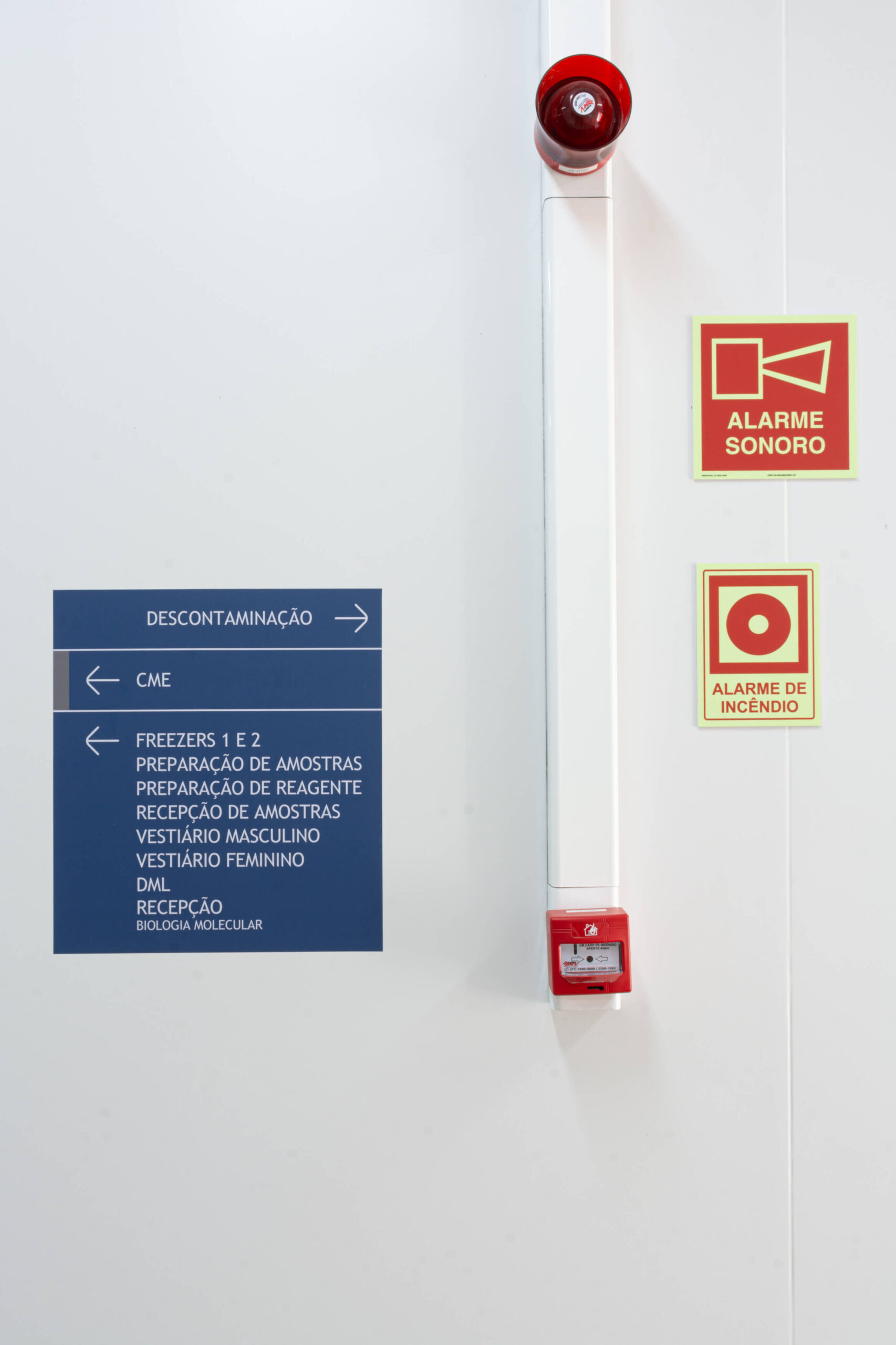 Parede branca com placas de sinalização interna, alarme sonoro vermelho e um aviso de 