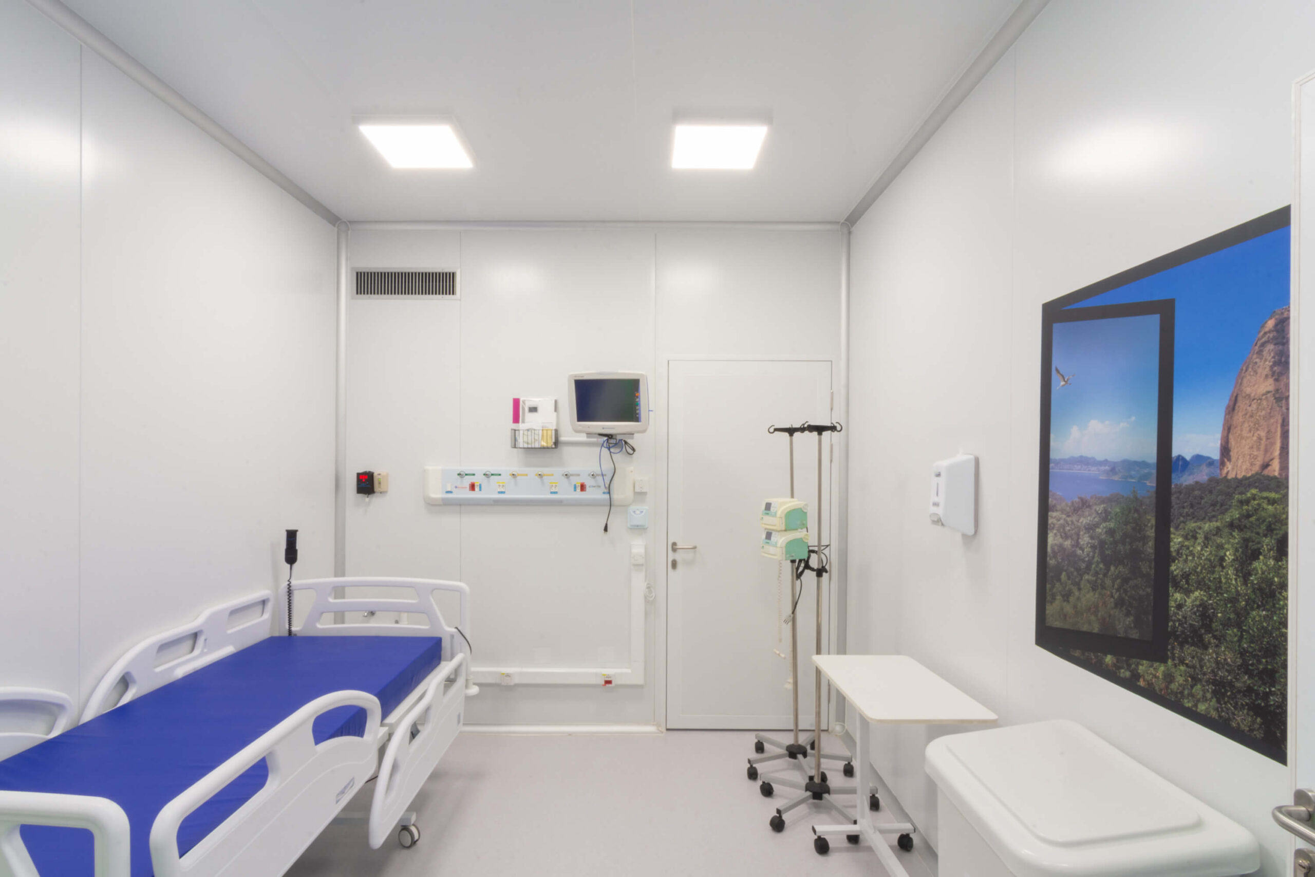 Interior de um quarto hospitalar humanizado e limpo, com cama branca e colchão azul, monitor de sinais vitais preso a parede junto com saídas de gás. Em outra parede, uma imagem que simula uma janela com vista para o Pão de Açúcar.