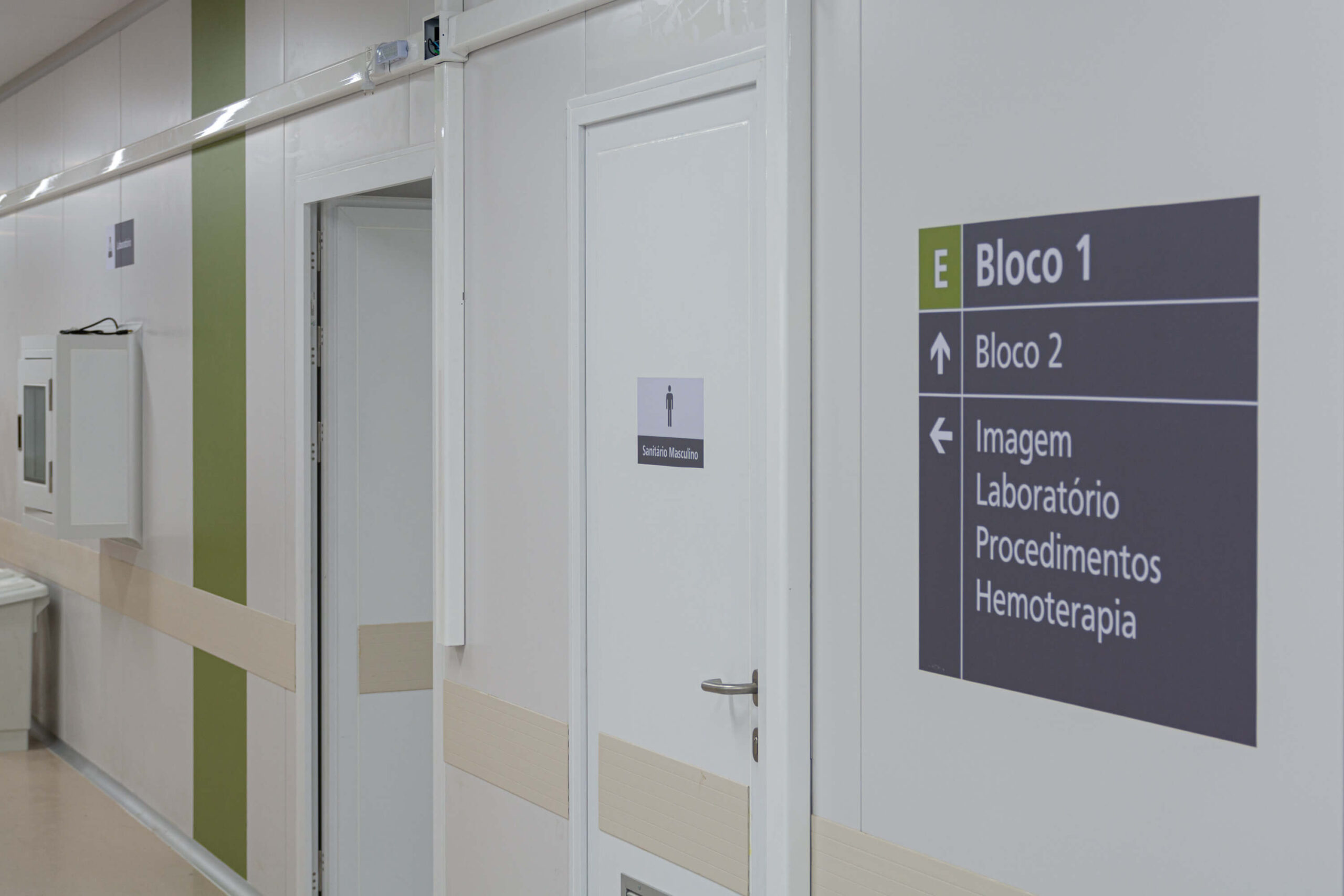 Corredor com paredes e portas na cor branca, placas de sinalização indicando ser o Bloco 1.