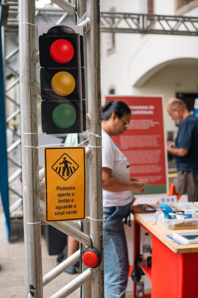 Close de detalhes da exposição, com a sinalização de semáforo e placa travessia com chamada para pedestres