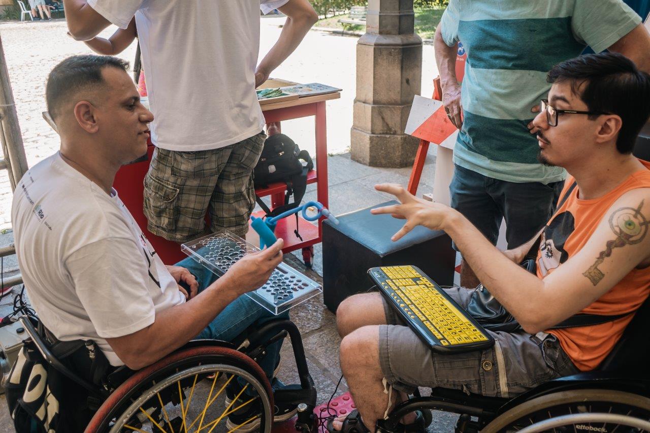 Dois homens em cadeiras de rodas conversam trocam sobre um teclado e objetos adaptados