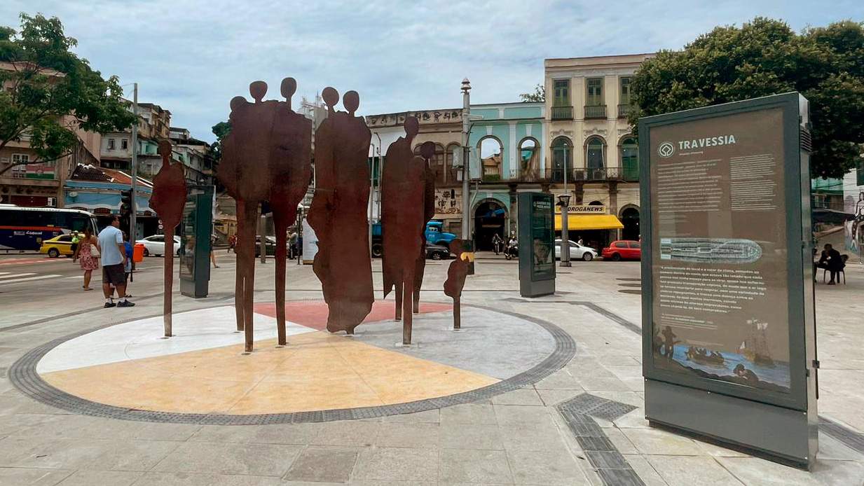 Instalação artística em uma praça pública, cercada por edifícios históricos de fachada colonial e árvores. No centro, sobre um piso circular dividido em quatro setores, erguem-se figuras humanas verticais, recortadas em chapas metálicas de aço cortem, com coloração avermelhada. As silhuetas representam pessoas negras, dispostas lados a lado em diferentes alturas, formando o mapa da África. Ao redor da escultura, painéis com textos dos módulos da exposição com diferentes cores de fundo.