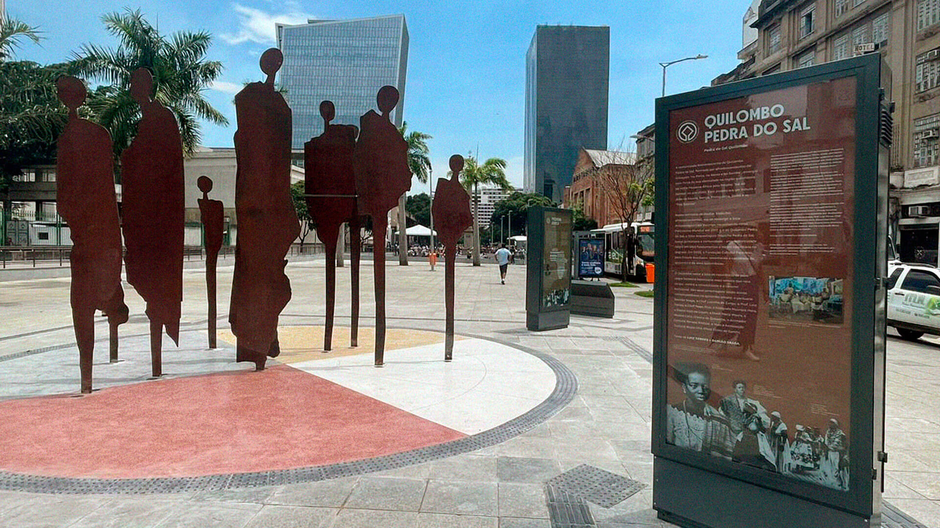 Instalação artística em uma praça pública, cercada por edifícios históricos. No centro, sobre um piso circular dividido em quatro setores, erguem-se figuras humanas verticais, recortadas em chapas metálicas de aço cortem, com coloração avermelhada. As silhuetas representam pessoas negras, dispostas lados a lado em diferentes alturas. Ao redor da escultura, painéis com textos dos módulos da exposição com diferentes cores de fundo.