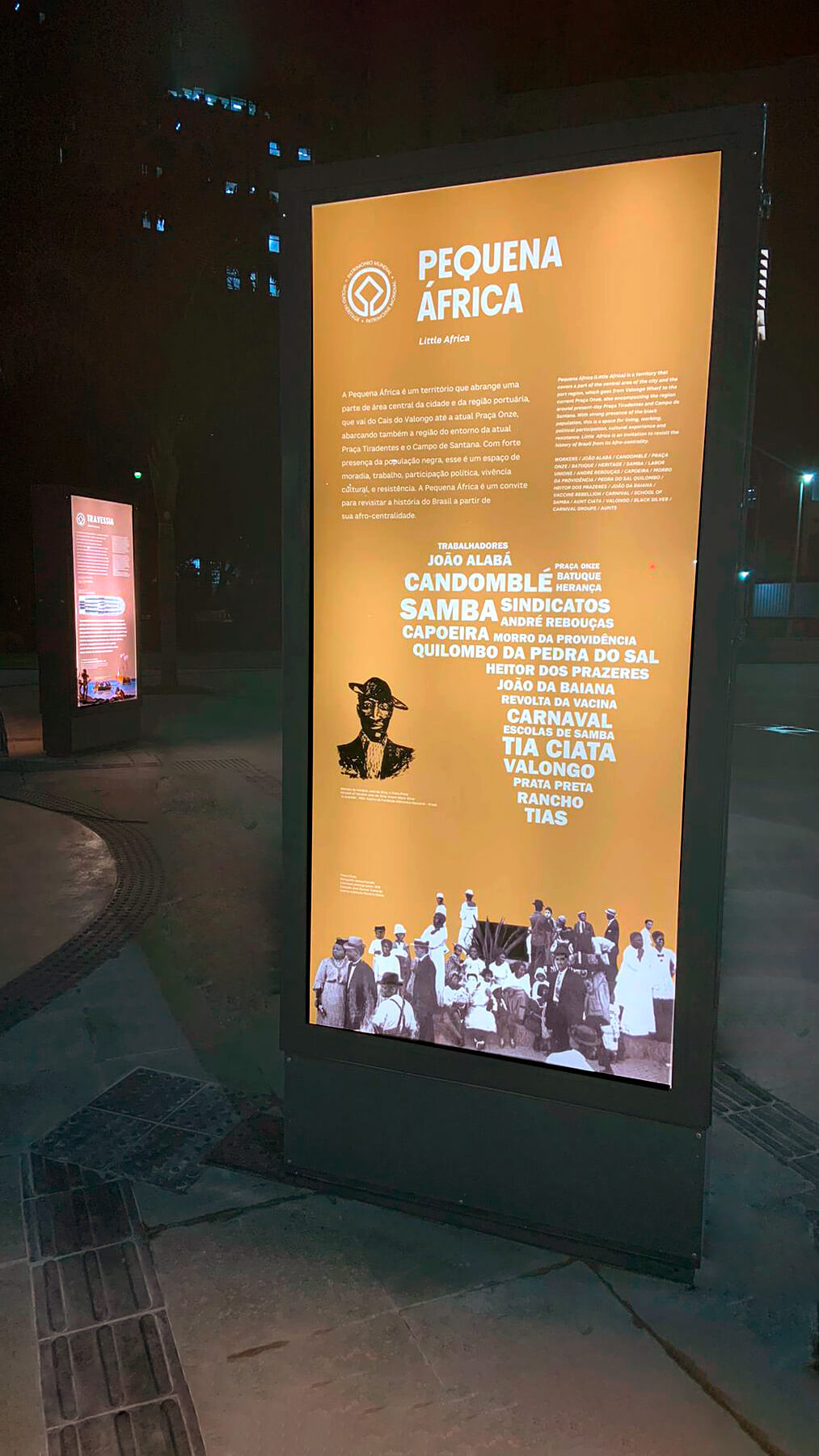 Painel de texto do módulo Pequena África, na exposição permanente do Cais do Valongo, possui cor amarela de fundo e recorte de imagens em preto e branco. Iluminado com luz de led interna, brilha na noite escura.
