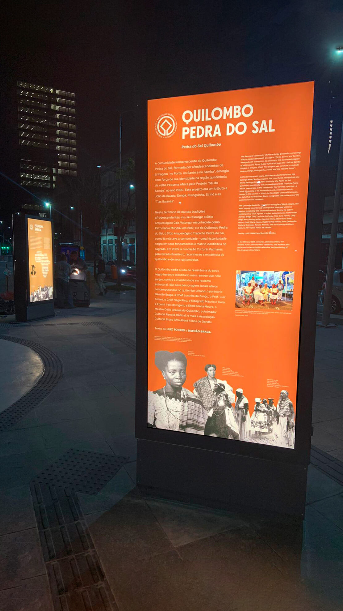 Painel de texto do módulo Quilombo Pedra do Sal, na exposição permanente do Cais do Valongo, possui cor laranja de fundo e recorte de imagens em preto e branco. Iluminado com luz de led interna, brilha na noite escura.