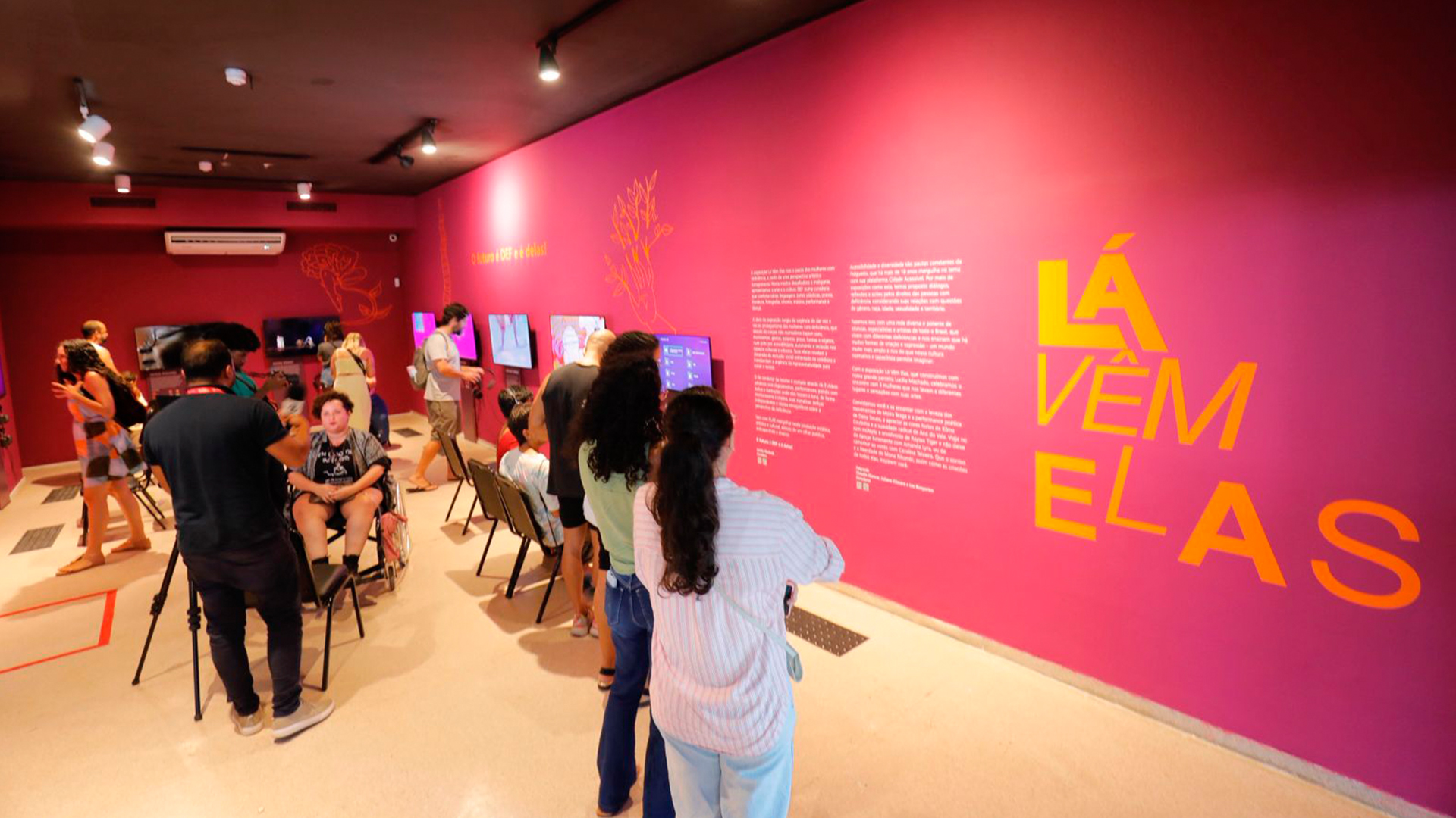 Sala de exposição com paredes em tom de magenta e textos em laranja. O texto principal na parede diz “Lá Vêm Elas”. Visitantes observam telas de vídeo fixadas na parede e algumas pessoas estão sentadas em cadeiras assistindo ao conteúdo.