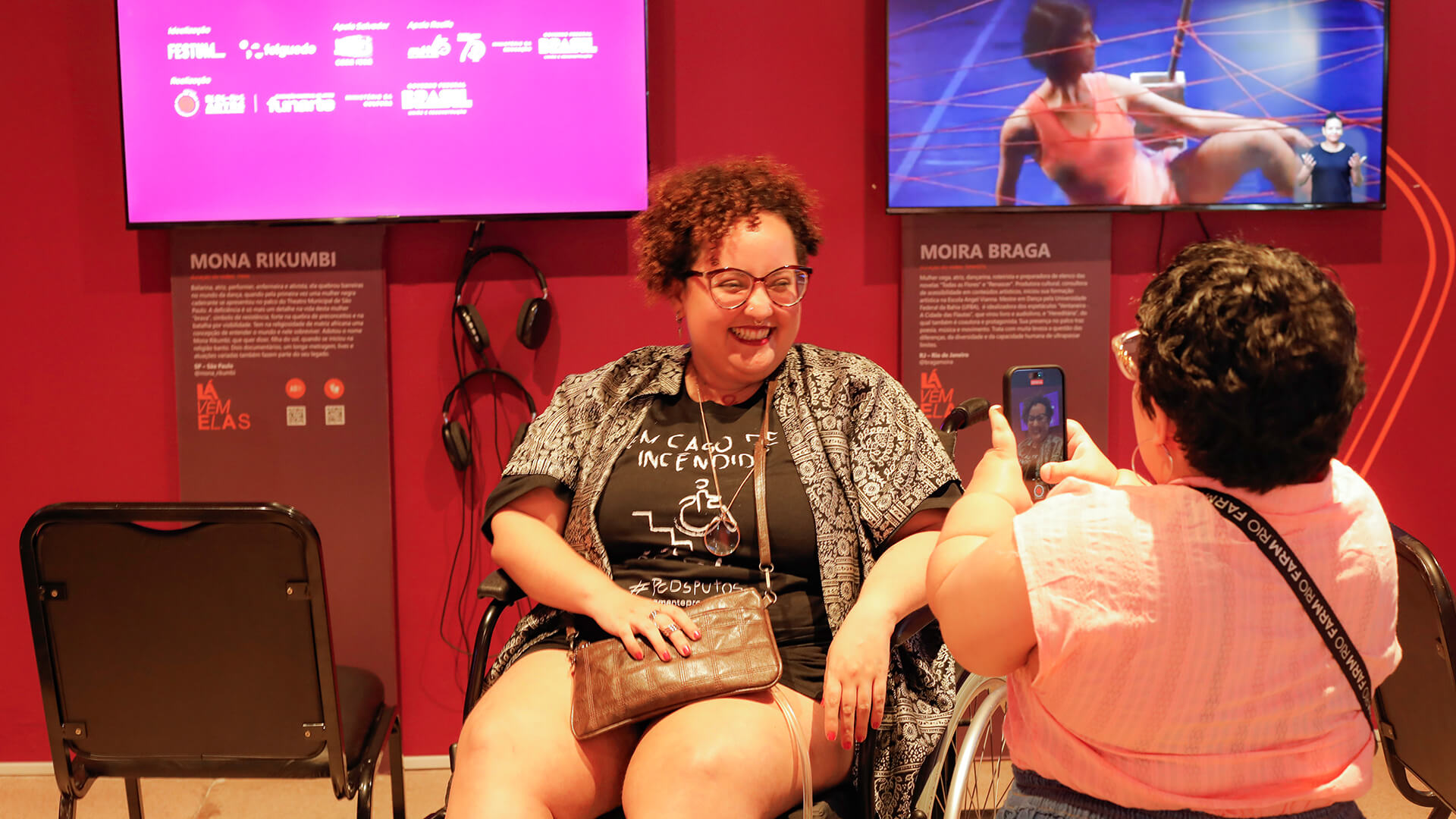 Duas mulheres com deficiência conversam e sorriem em frente a uma parede rosa de exposição. Uma delas, de cabelo curto e cacheado, usa cadeira de rodas e camiseta preta e um kimono estampado. A outra mulher está de costas, tirando uma foto com o celular. Atrás delas, há telas exibindo vídeos e textos explicativos com os nomes “Mona Rikumbi” e “Moira Braga”.