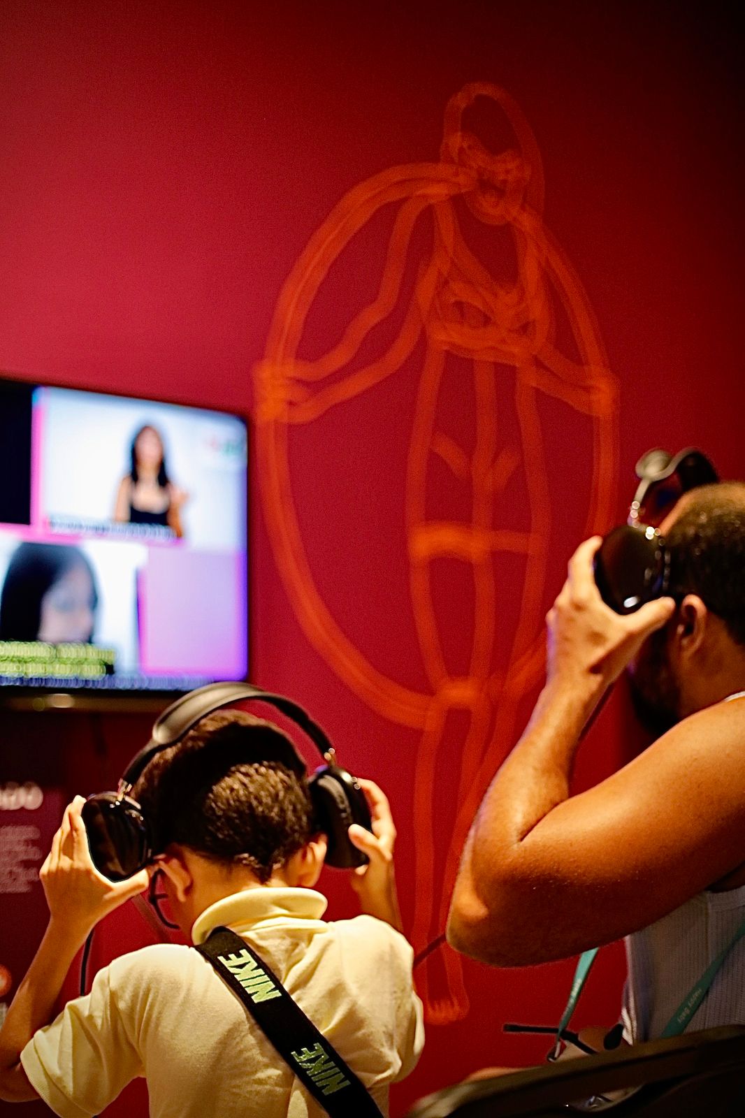 Um homem e um menino colocando fones de ouvido para assistir a vídeo em televisão. Ao fundo uma parede magenta com ilustração de uma mulher segurando um bambolê em suas costas.