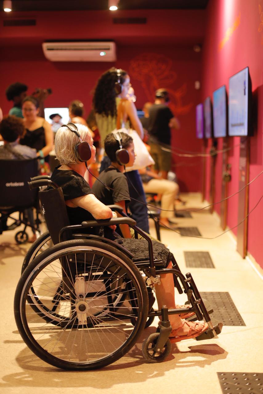 Mulher idosa de cabelos grisalhos em cadeira de rodas e um menino ao lado, ambos usando fones de ouvido, assistem a um vídeo em televisão. Outras pessoas, algumas também em cadeiras de rodas, observam as telas ao redor na mesma sala.