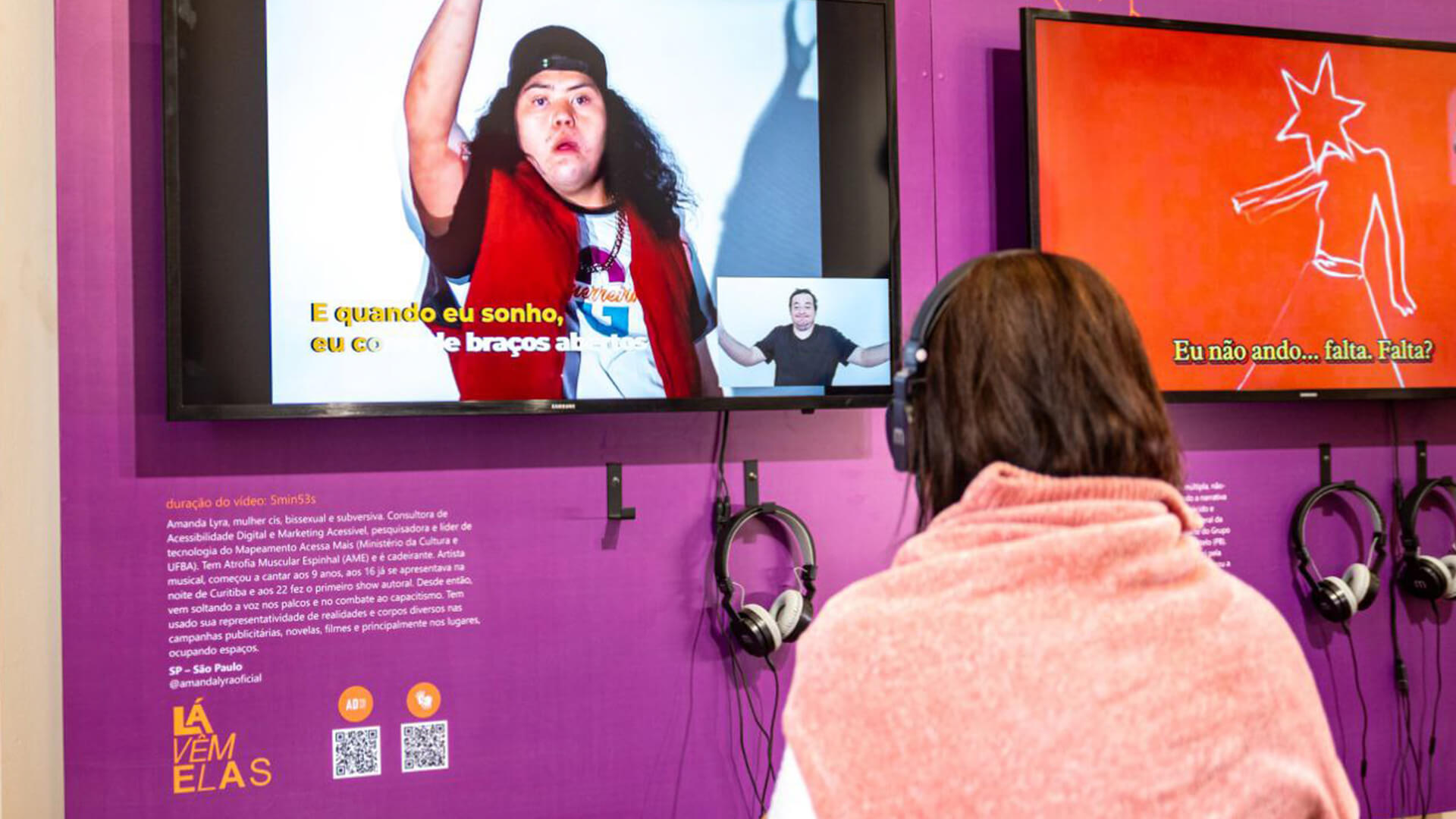 Mulher sentada de costa para a foto e de frente para um painel roxo com texto descritivo e uma grande televisão conectada com fones de ouvido.