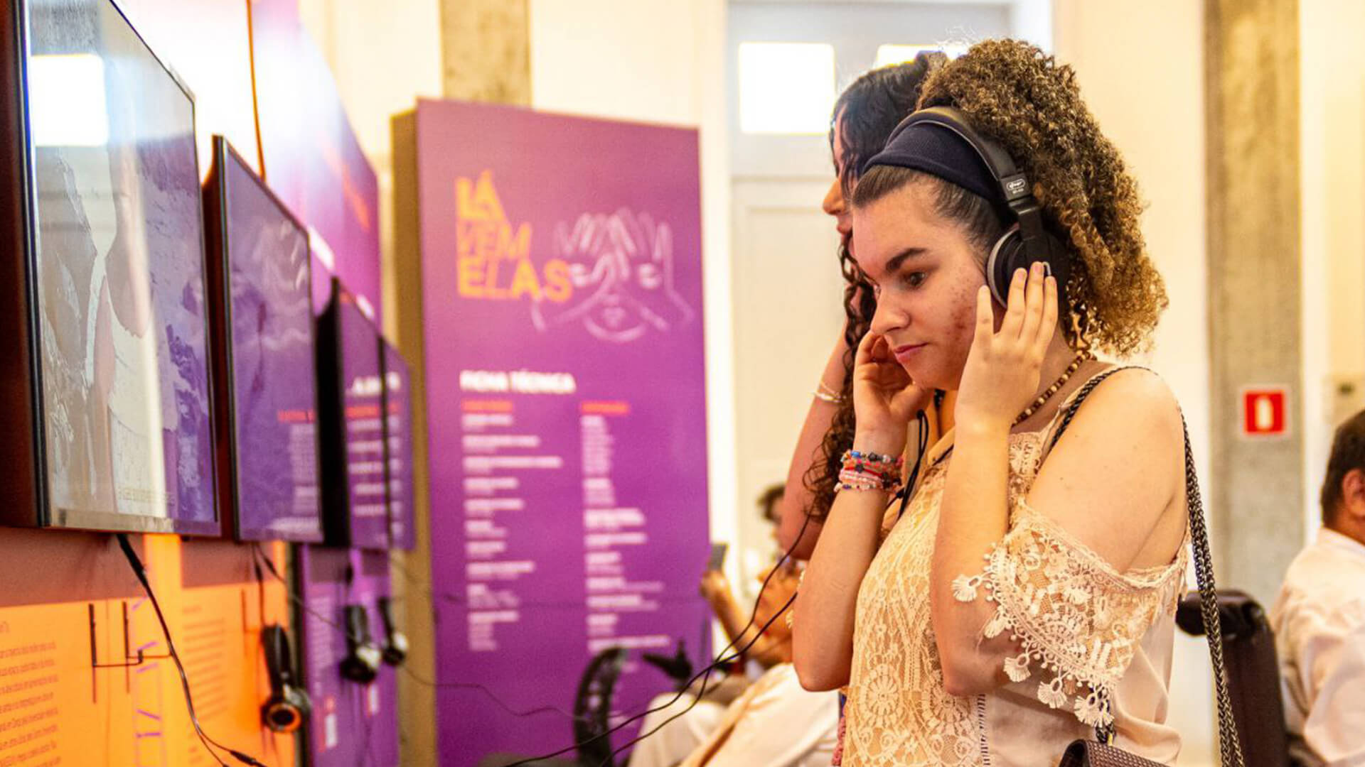Mulher com fones de ouvido conectado em TV, ouvindo áudios da exposição. Painéis de conteúdo nas cores roxo e laranja ao redor da sala.