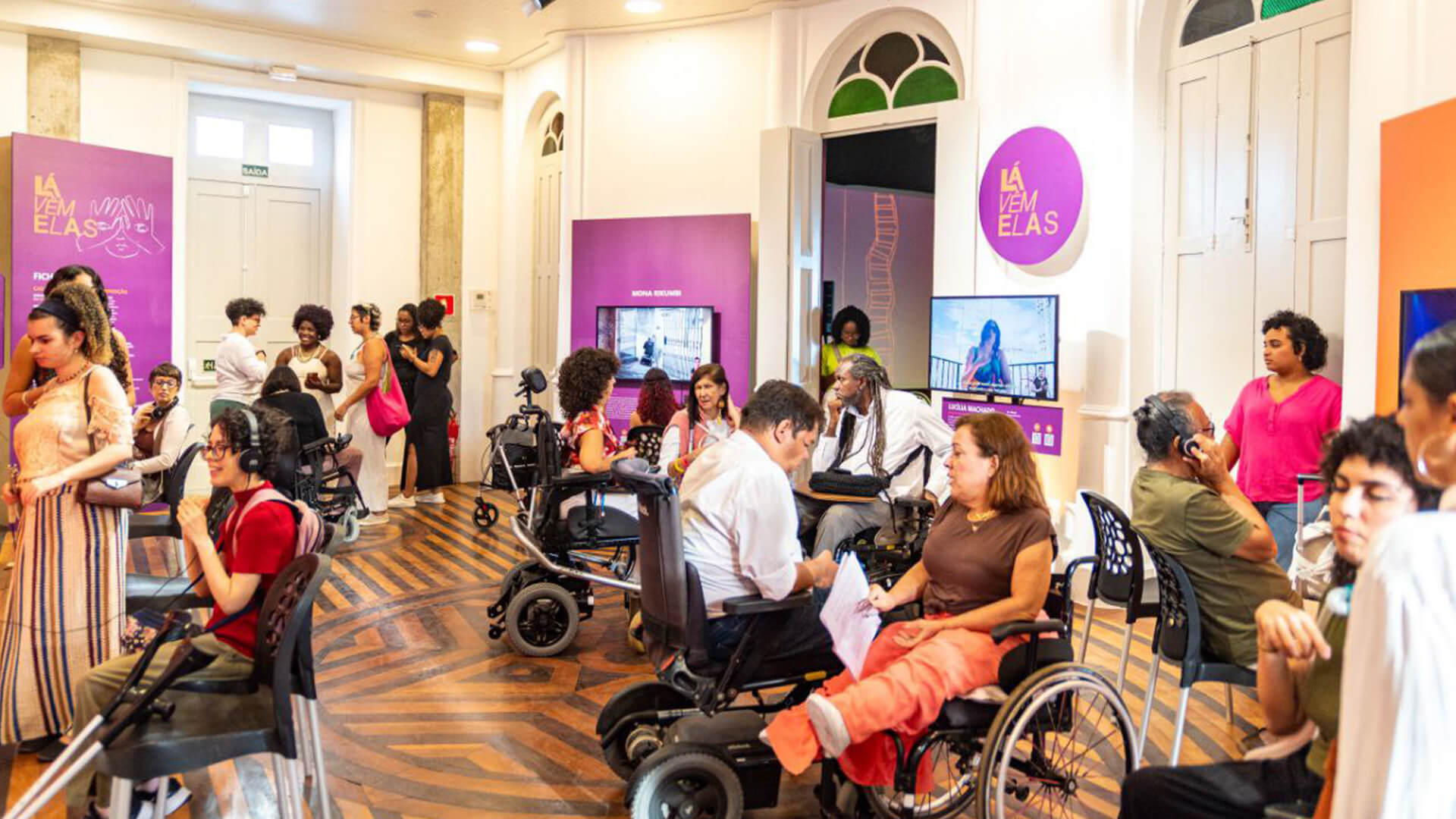 Grupo de pessoas diversas juntas em uma sala. Ao centro, um grupo de pessoas com deficiência em cadeiras de rodas, conversando e interagindo com a exposição. O espaço interno com paredes brancas, piso de madeira com dois tons de marrom e painéis roxos com ilustrações e textos na cor laranja, algumas televisões e fones de ouvidos.