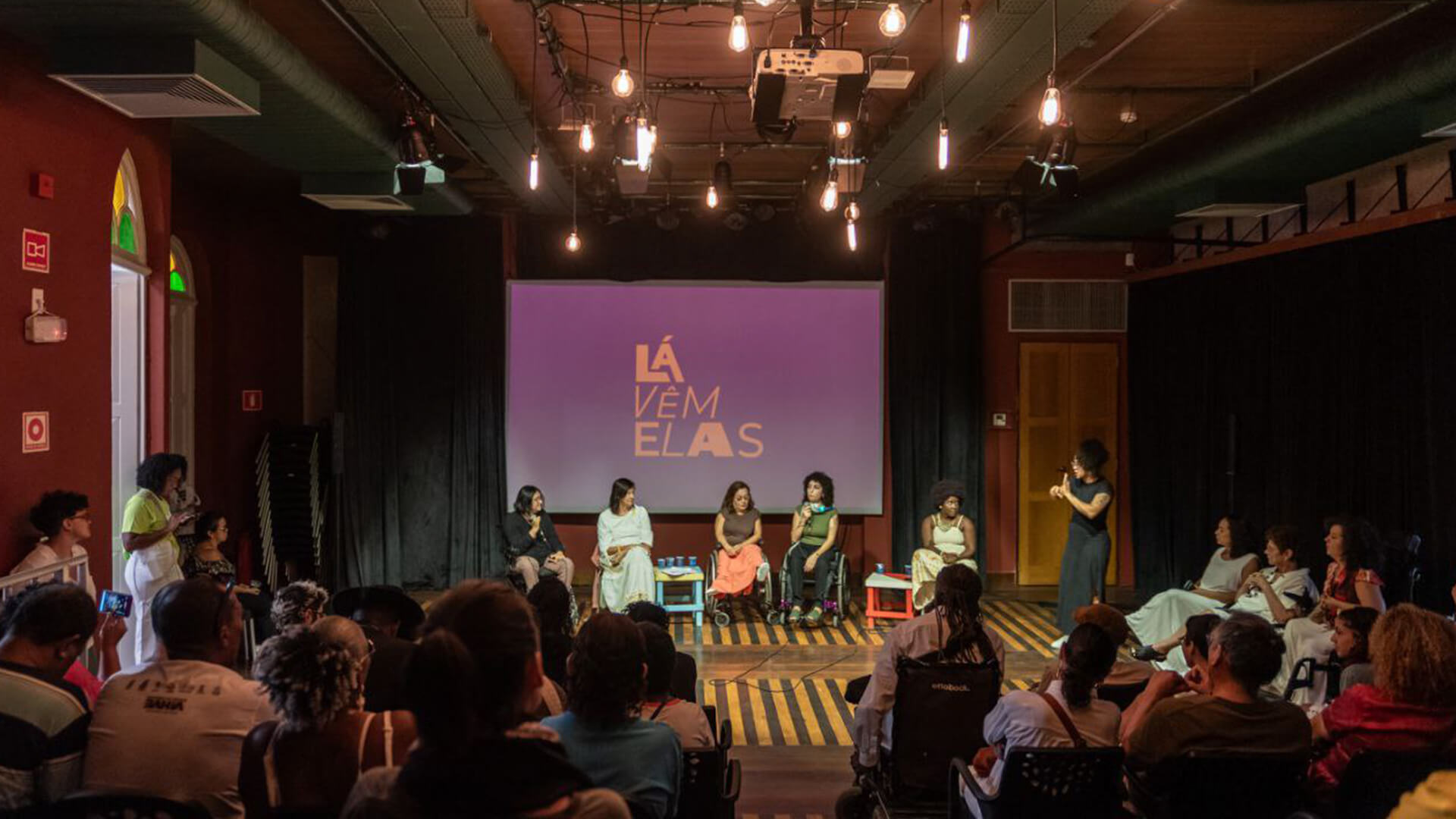 Sala amplas com lâmpadas penduradas no teto fazendo a iluminação do ambiente, plateia sentada ao redor da sala. Grupo de mulheres com deficiências reunidas em frente a telão projetando a logo “Lá vêm elas”.