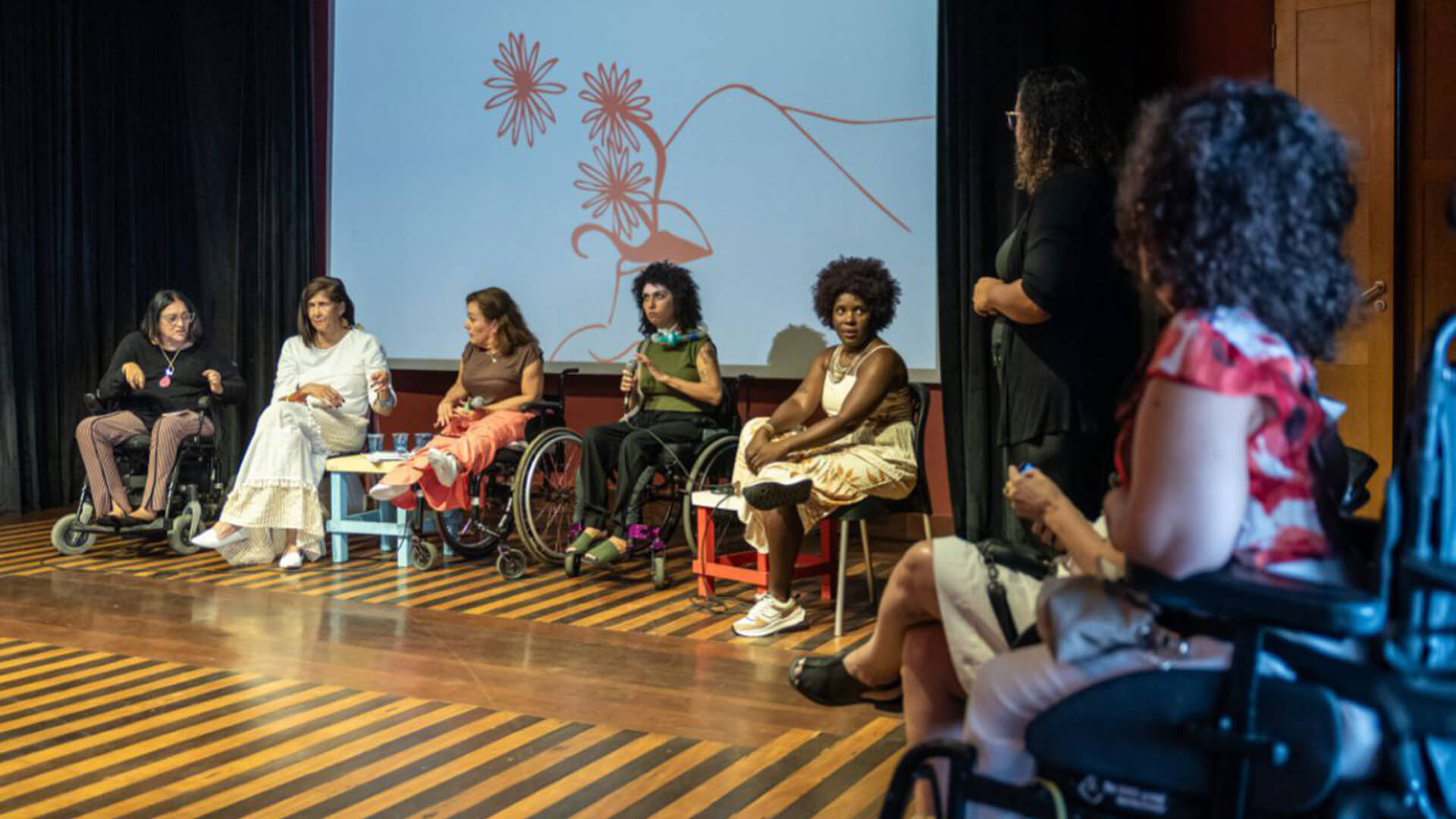 Grupos de seis mulheres reunidas em frente a um telão projetando uma ilustração de flores saindo de dentro de uma boca feminina. Quatro das mulheres reunidas estão em cadeira de rodas.