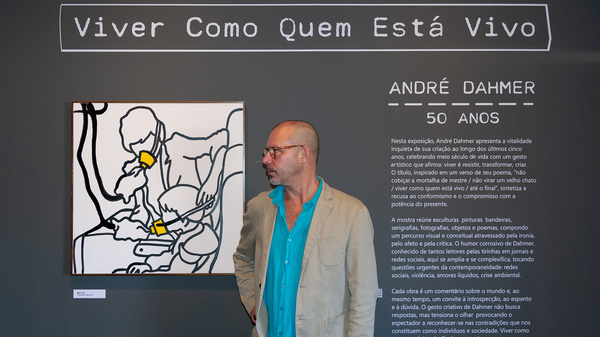 O artista Andr&eacute; Dahmer posa para foto, &eacute; um homem branco vestido uma camisa azul claro vibrante por baixo de um blazer bege, usa &oacute;culos. Dahmer est&aacute; centralizado em uma parede preta, entre um texto explicativo da exposi&ccedil;&atilde;o e um quadro branco com uma ilustra&ccedil;&atilde;o feito de linhas pretas de uma pessoa colocando m&aacute;scaras de oxig&ecirc;nio em uma crian&ccedil;a. Sob ele o t&iacute;tulo &ldquo;Viver como quem est&aacute; vivo&rdquo;