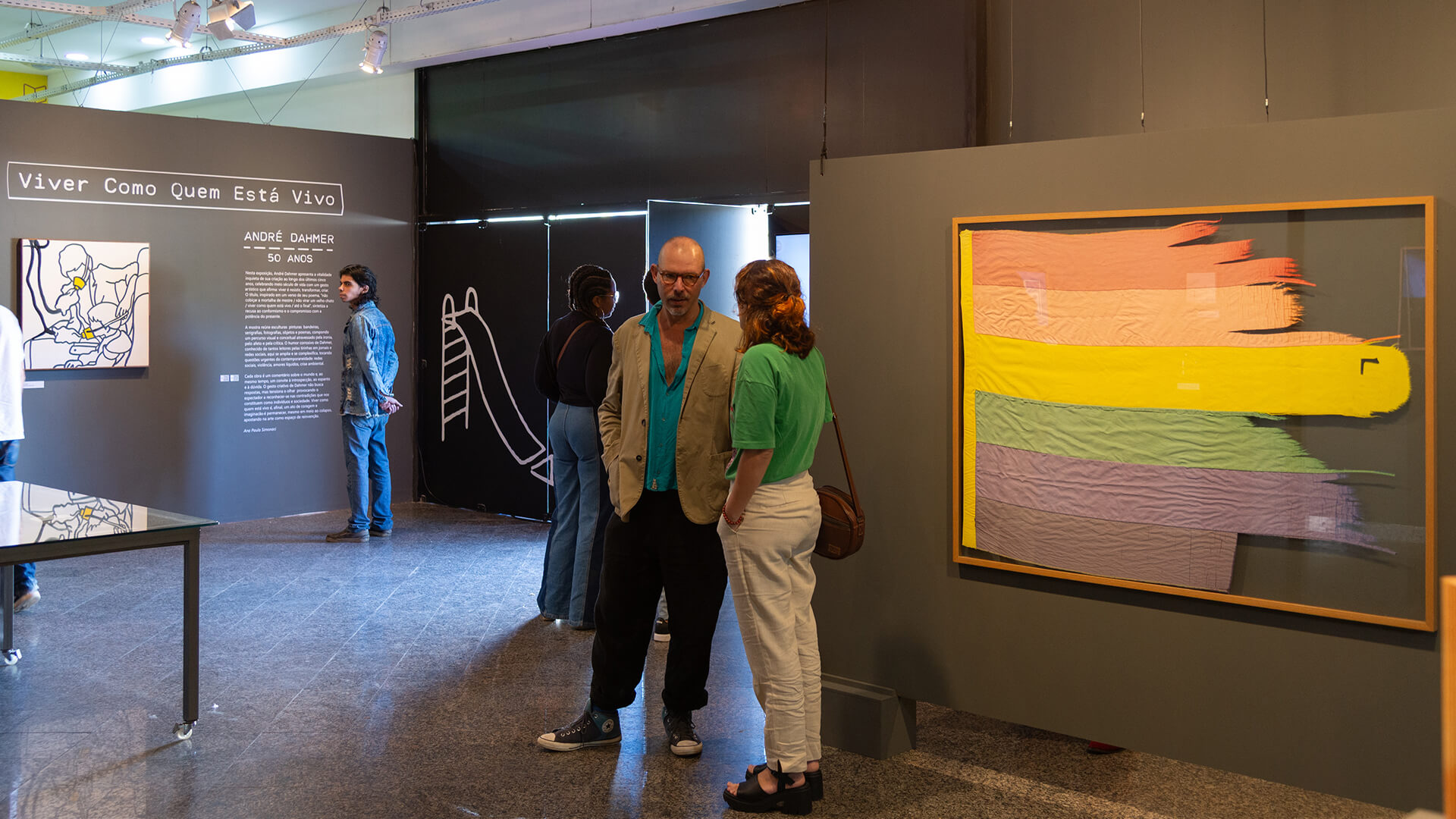 Sala expositiva com parede na cor preta, na lateral direita em destaque, uma bandeira com as cores do a arco-iris 