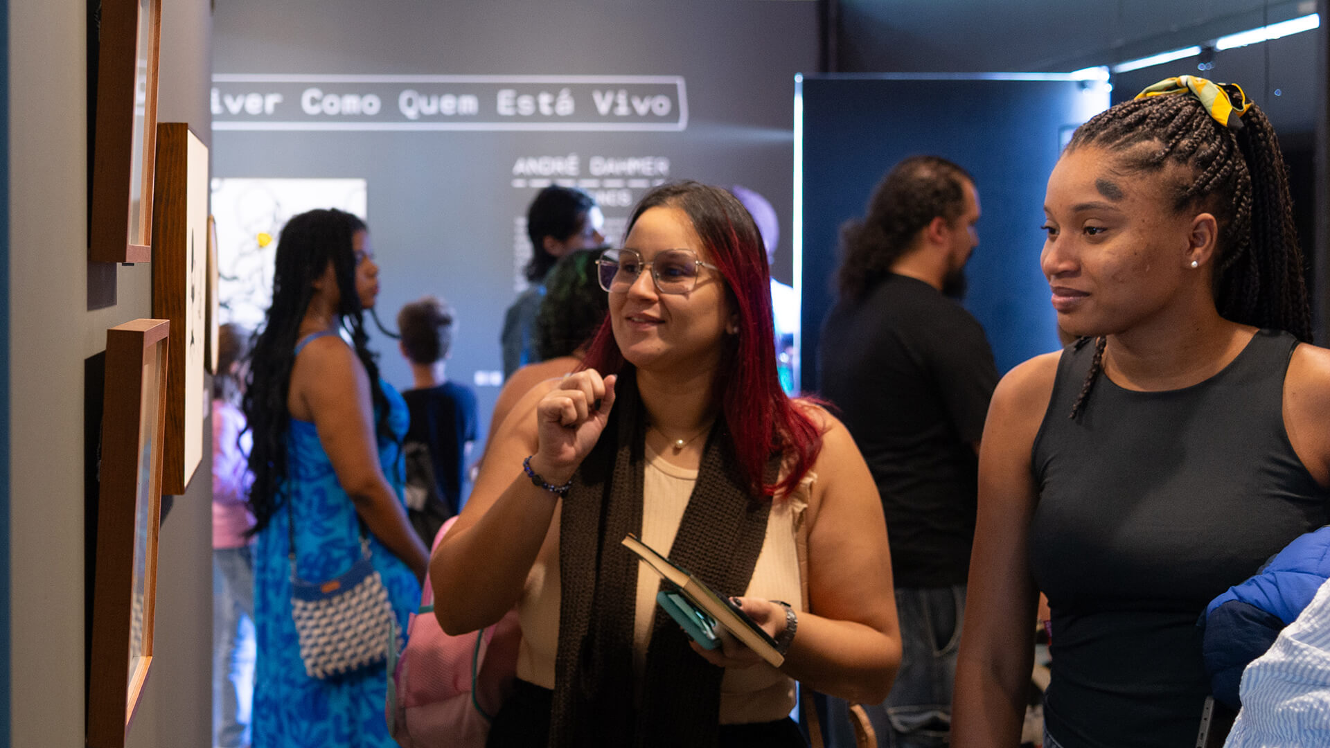 Duas mulheres, lado a lado, observam uma parede de quadros. 