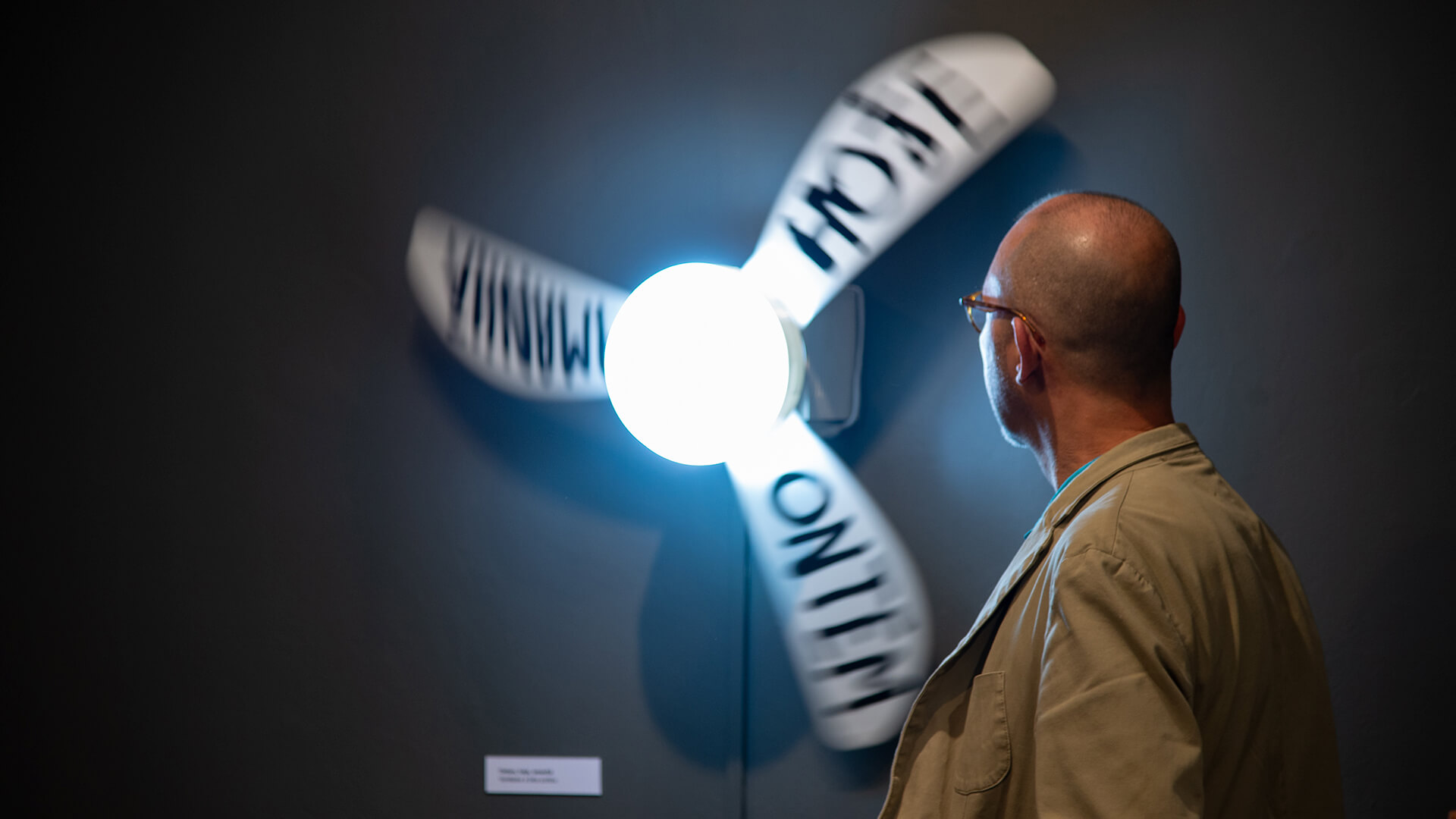 Fotografia da exposiçãHomem branco de lado observando ventilador de teto branco, preso em uma parede, com sua luz acesa e com palavras escritas em cada uma das suas pás “ontem” “hoje” “amanhã”.